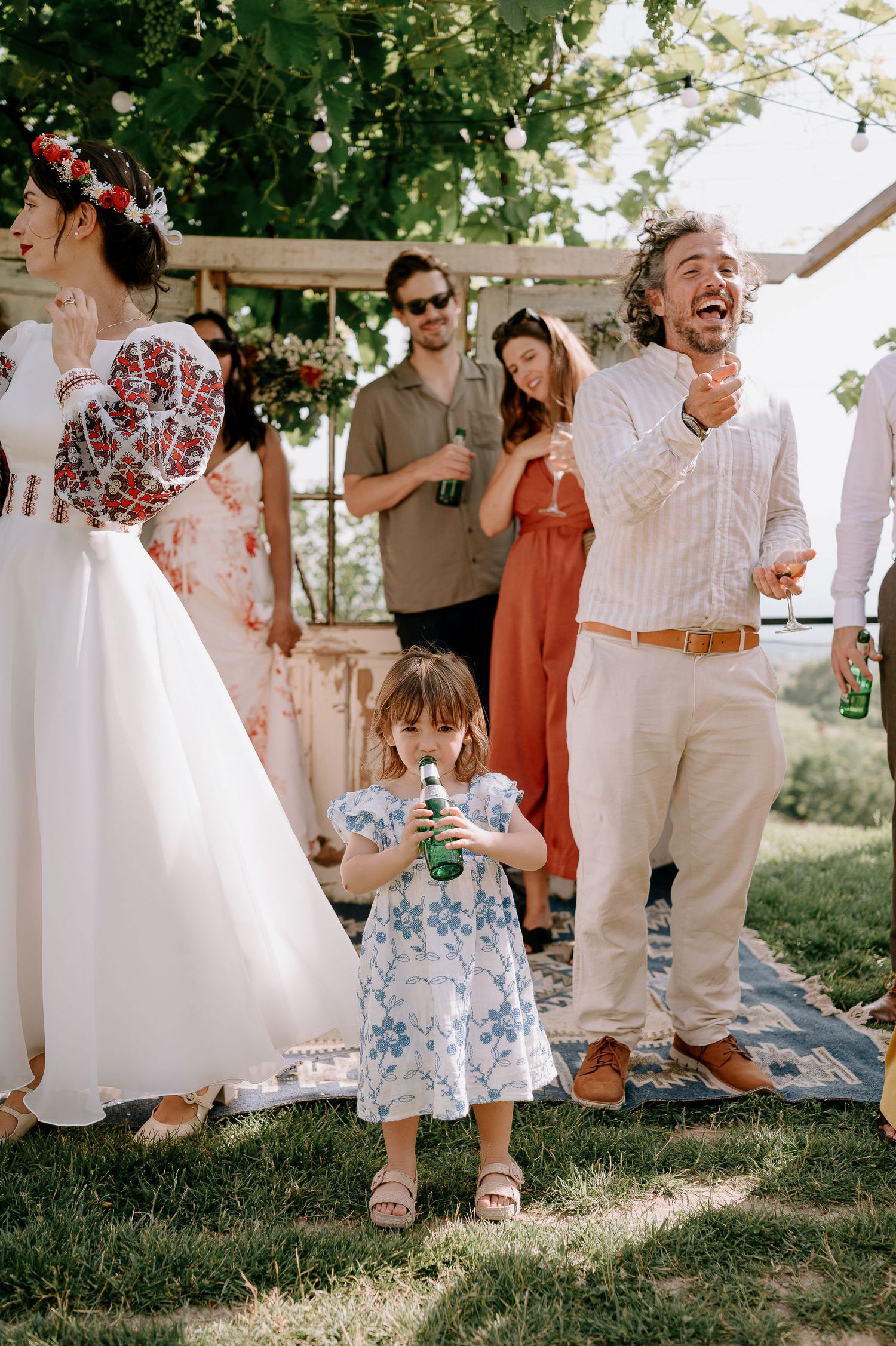 A young child in a floral dress enjoys a drink during an outdoor wedding celebration.