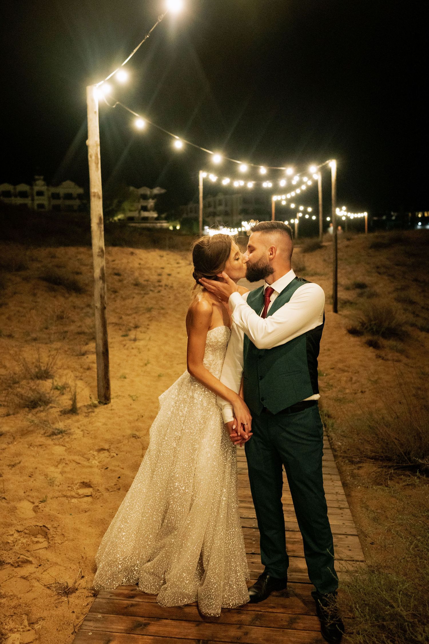 A romantic nighttime wedding portrait beneath glowing string lights on a wooden walkway.