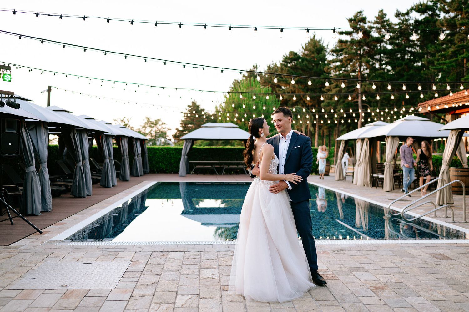 Elegant poolside wedding venue with string lights and stone patio during sunset.