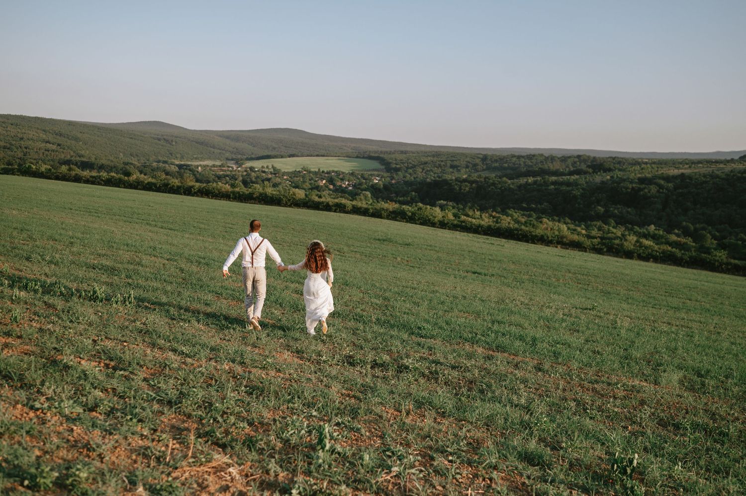 A couple in white wedding attire walks hand in hand across a scenic hilltop meadow.