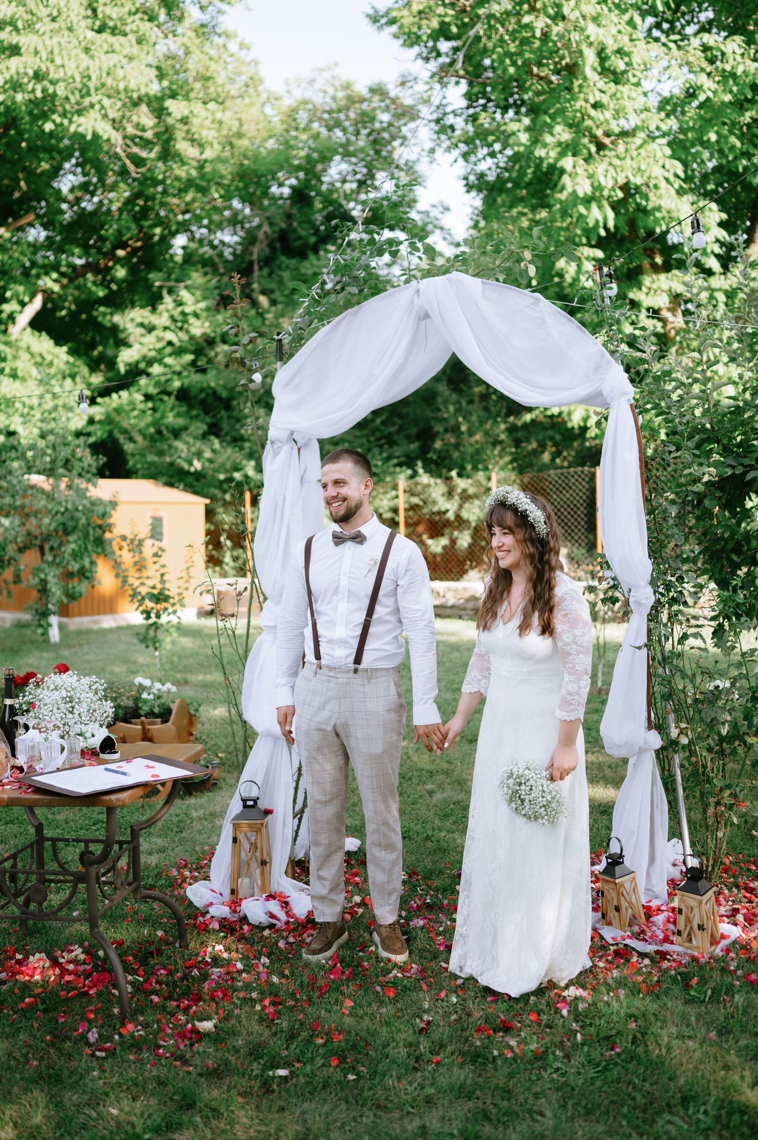 A garden wedding ceremony with white fabric arch and red flower petals scattered on the ground.