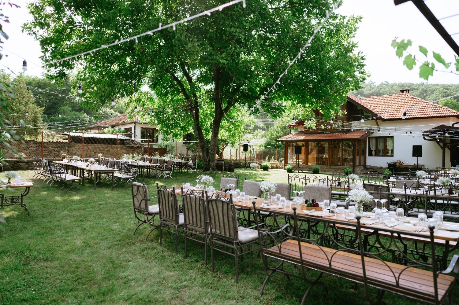 An outdoor wedding reception setup with white tables and chairs under string lights and trees.