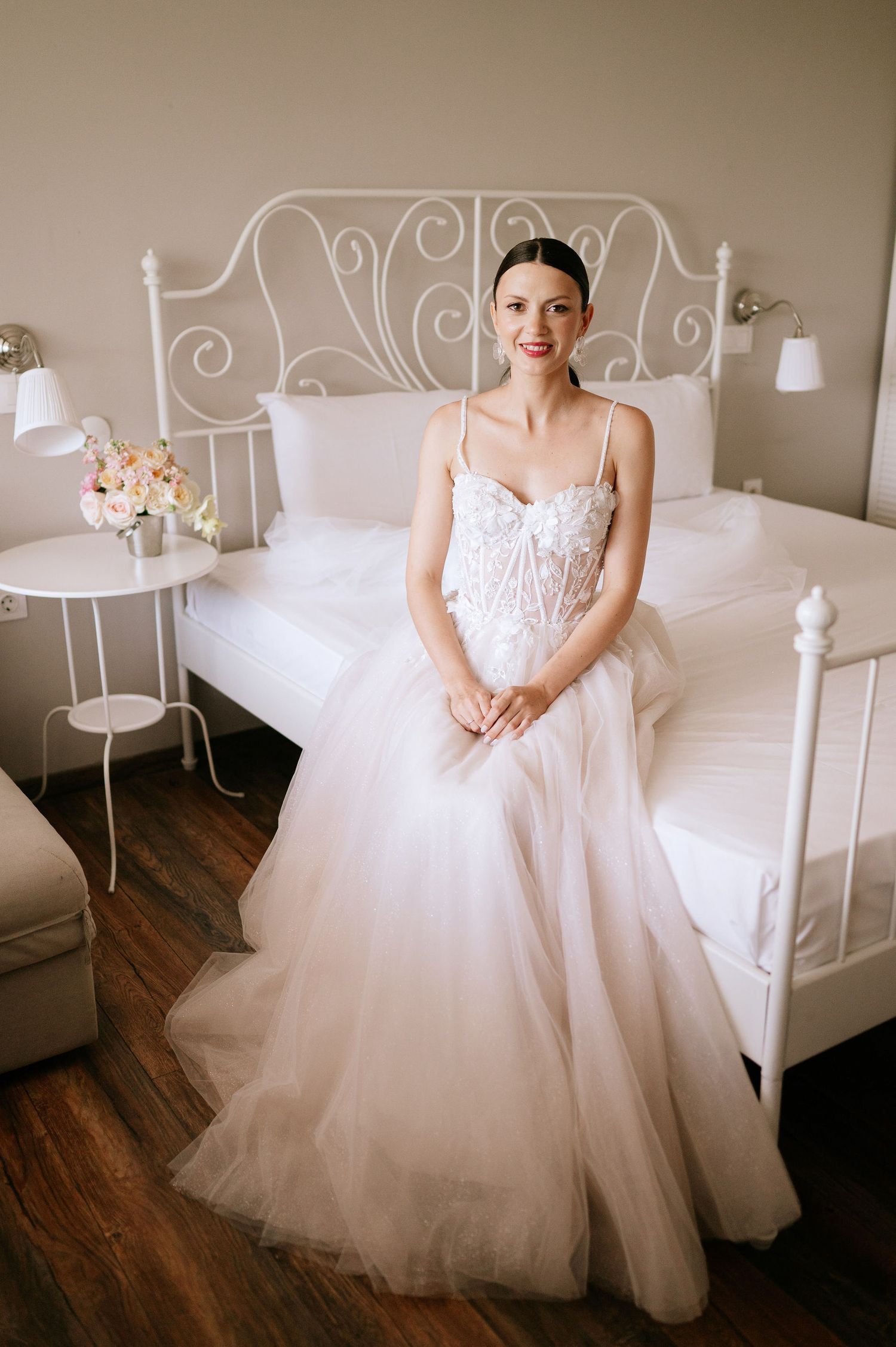 Romantic wedding dress with tulle skirt displayed against white iron bed frame in natural light.