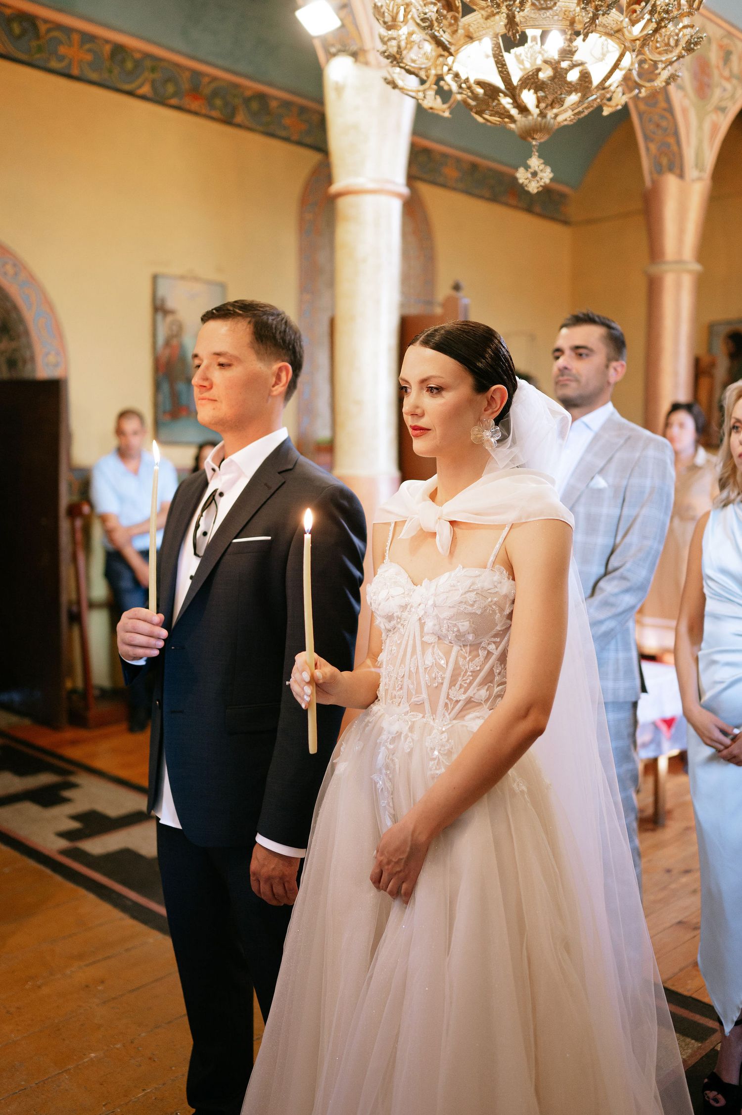 Wedding ceremony participants stand in formal attire holding candles in an elegant church setting.