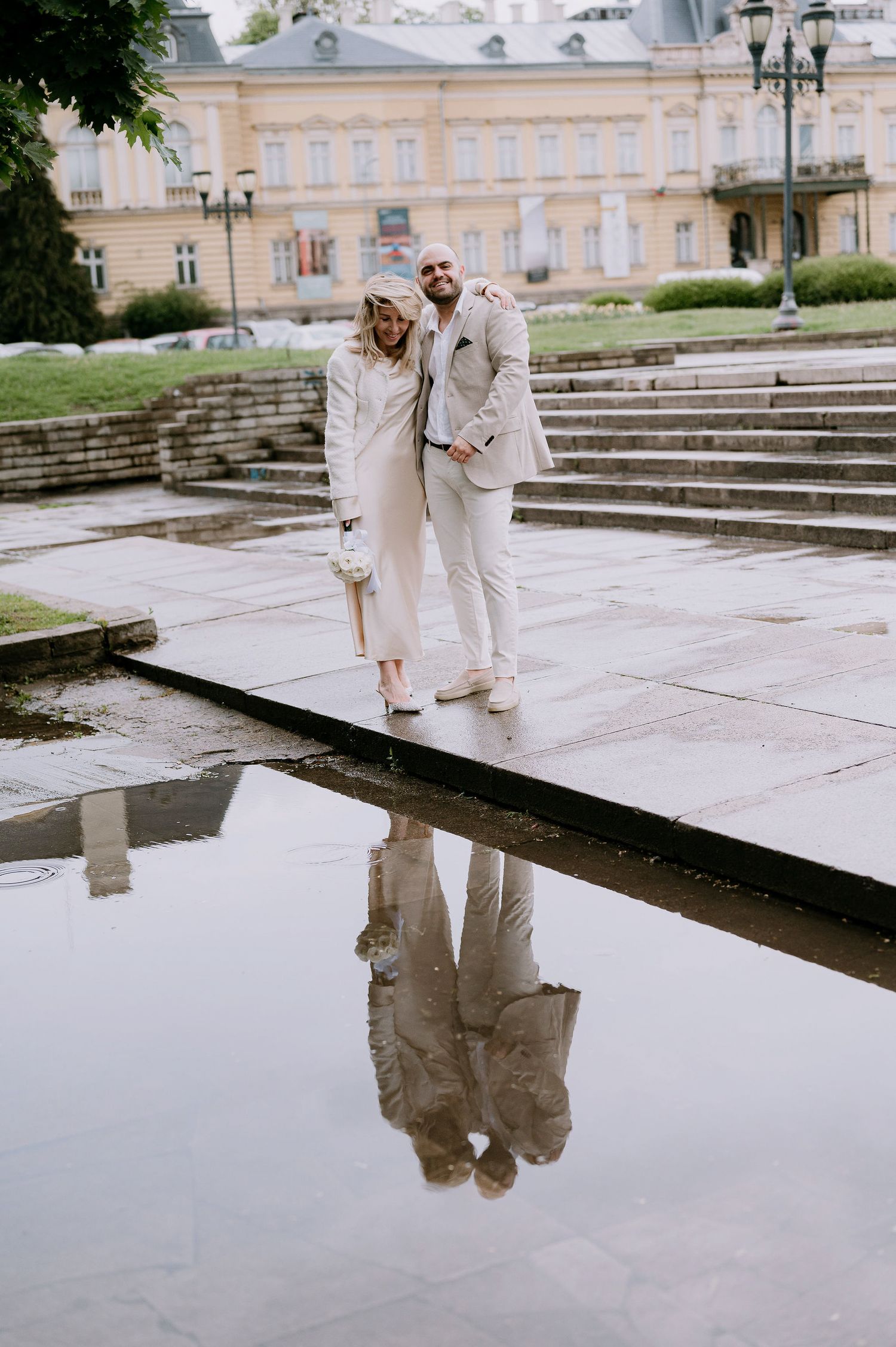 A couple in white outfits poses together on stone steps with reflective puddles near a historic building.
