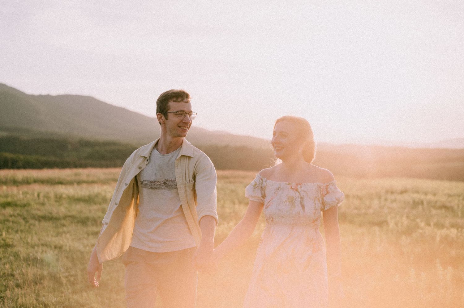 A couple walking together in a golden field at sunset with mountains in the background.