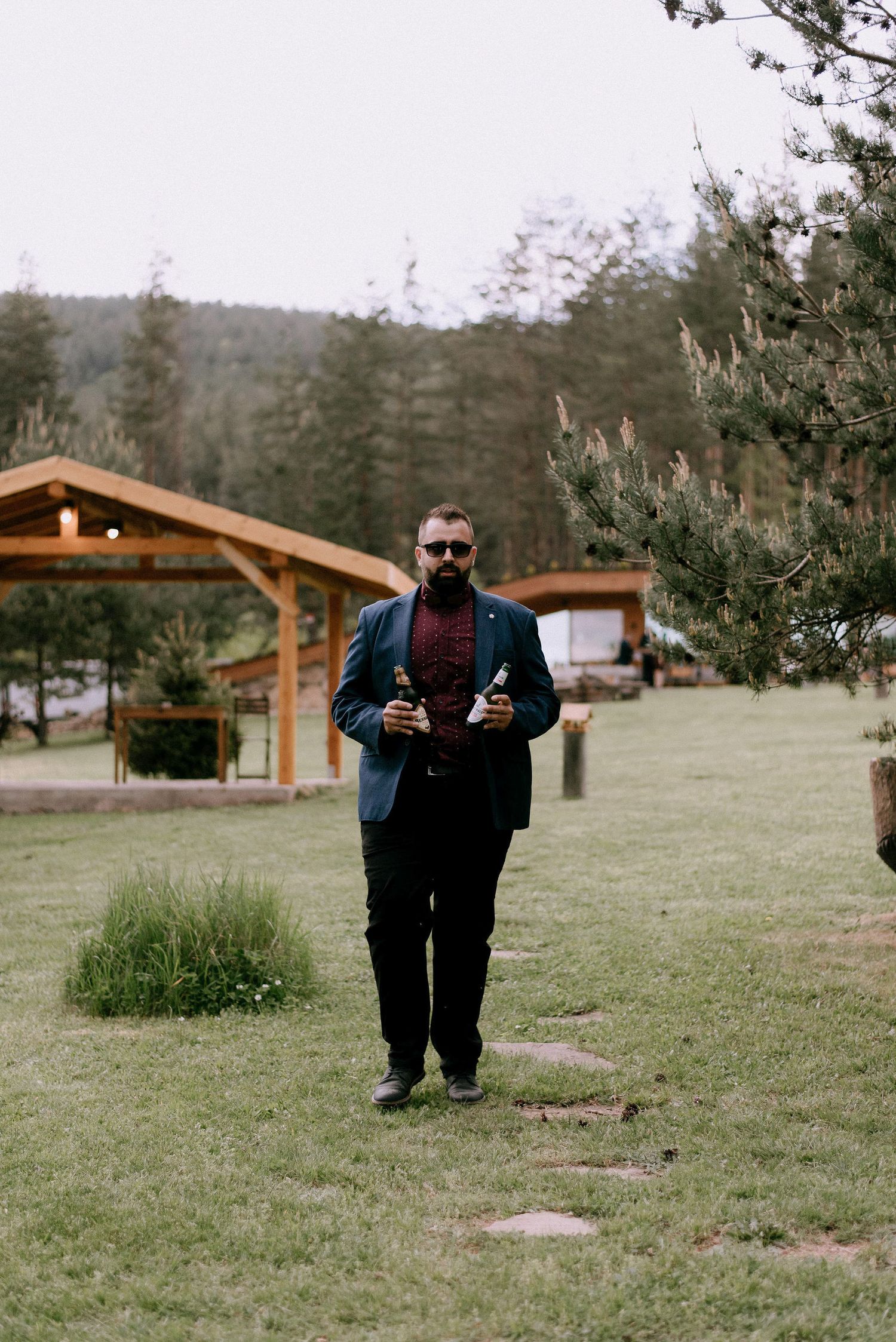 A person in formal attire poses with drinks on a grassy lawn with wooden cabins in the background.