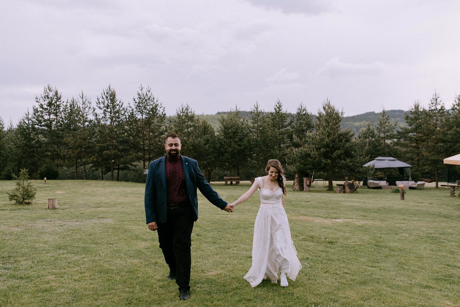 A couple walks hand in hand across a grassy field with pine trees and cabins in the background.