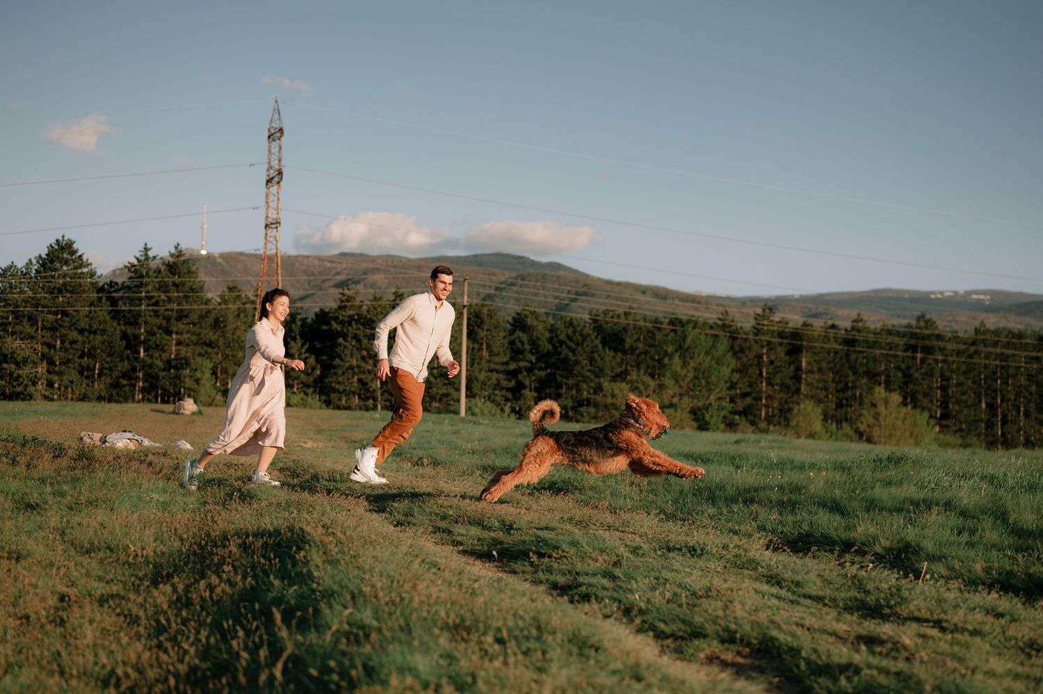 People and pets gather in a scenic mountain field during golden hour.
