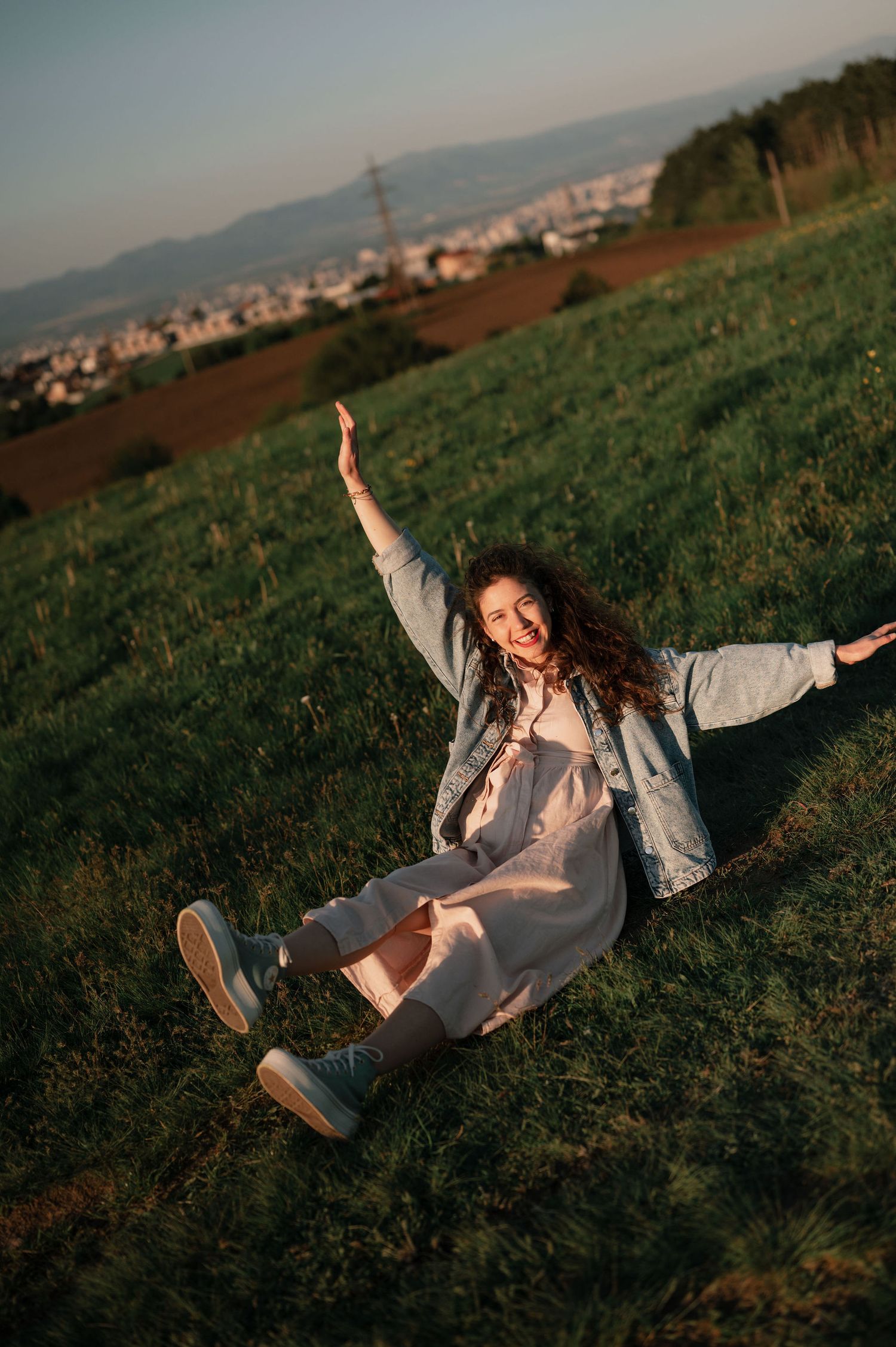 A person in a gray outfit lies joyfully on a grassy hillside overlooking a distant town during golden hour.