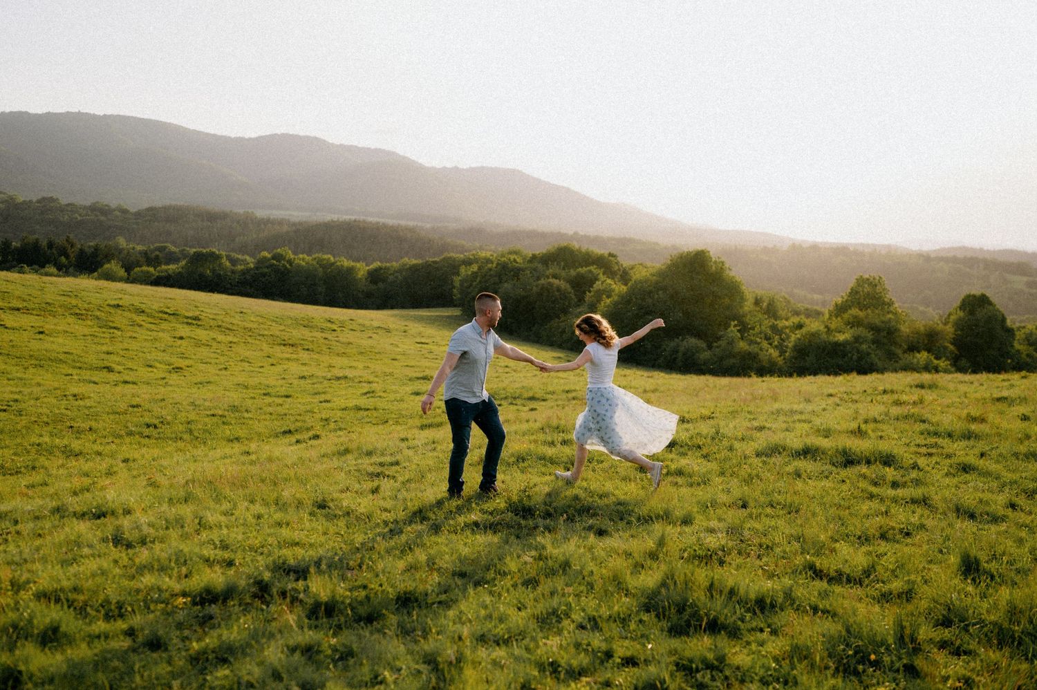 Playful couple runs through a grassy meadow with mountains on the horizon.