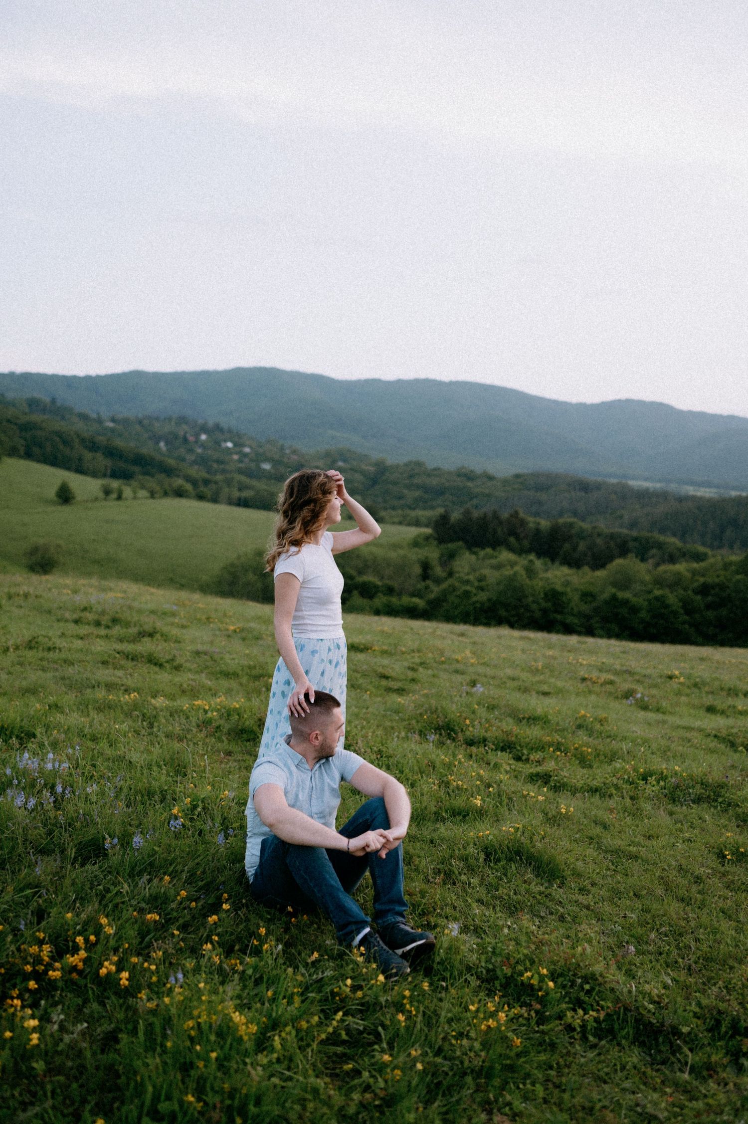 Two people relax in a grassy field with rolling mountains in the background.