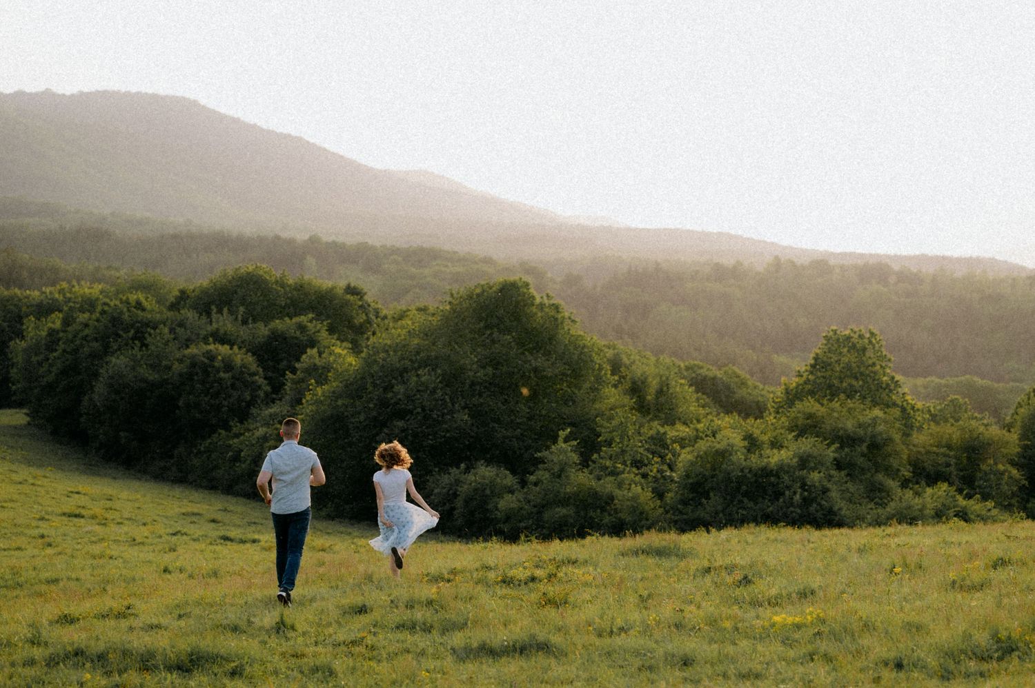 Two people walk together across a scenic hillside meadow at sunset with mountains and trees in the background.