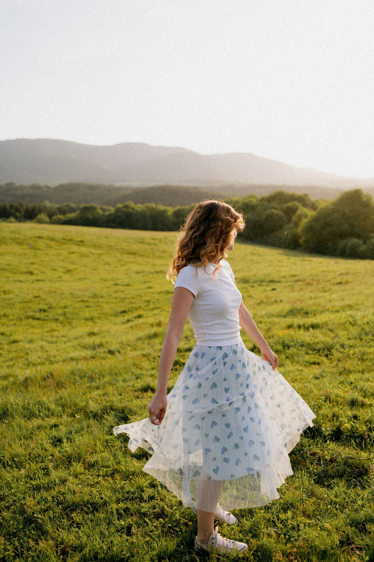 Dancing figure in a white floral dress spins joyfully across a green hillside at sunset.