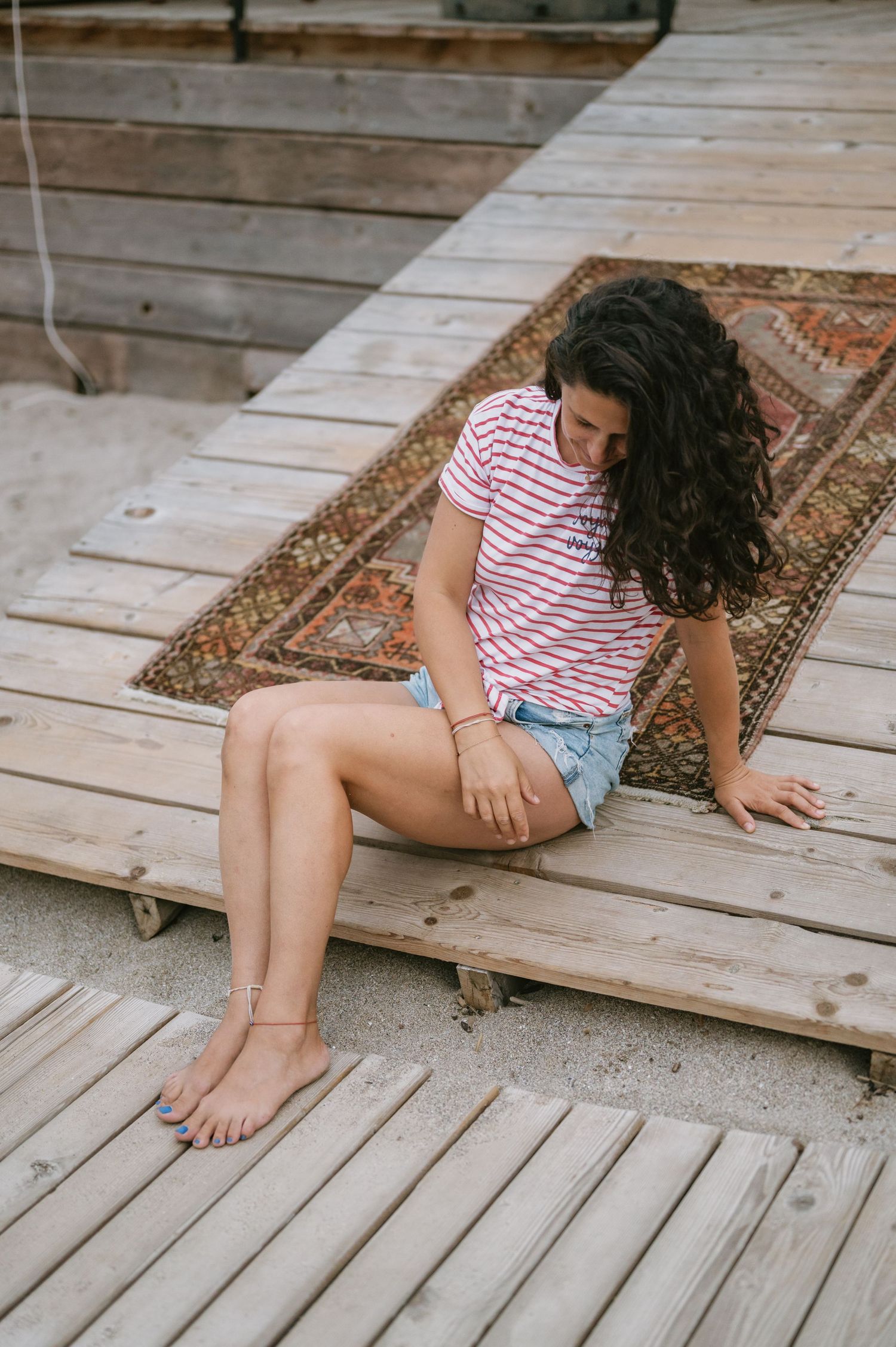 Young woman in striped tee and denim shorts sitting barefoot on wooden deck with vintage rug.