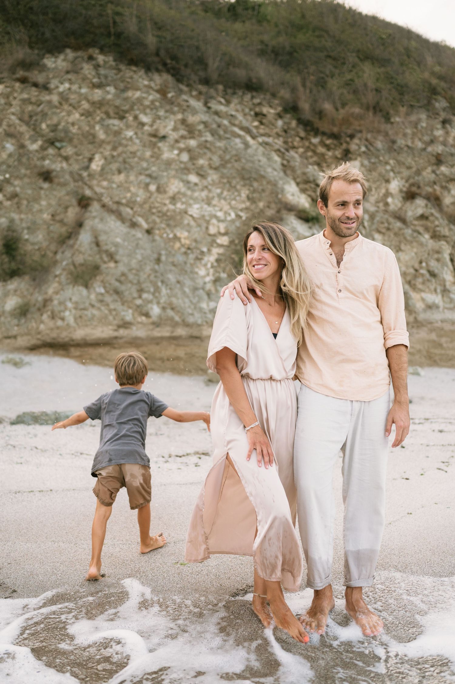 A happy couple walks barefoot on a sandy beach while their young child plays in the ocean waves near rocky cliffs.