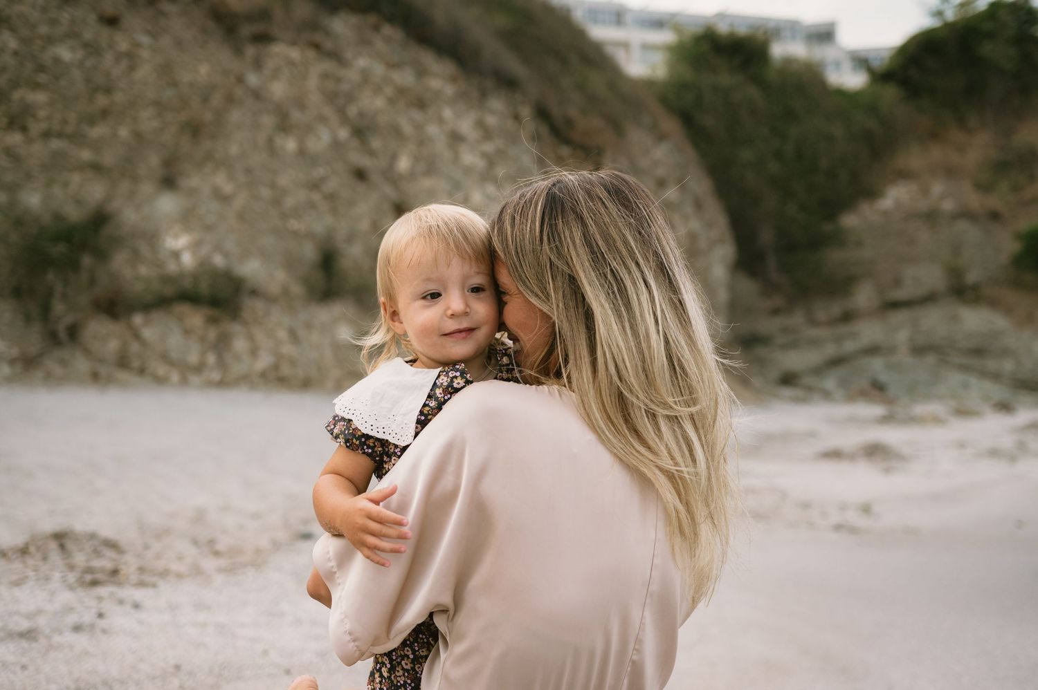 A mother tenderly kisses her smiling toddler on a beach with rocky cliffs in the background.