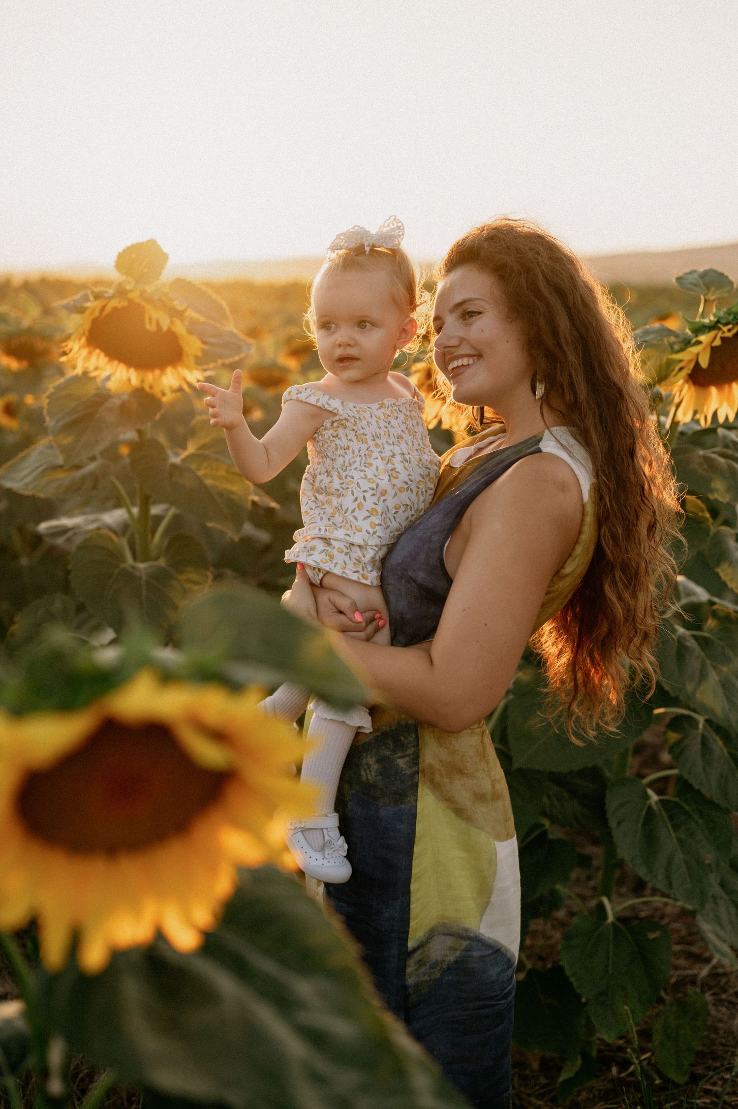 A smiling mother holds her baby girl in a golden sunflower field at sunset.