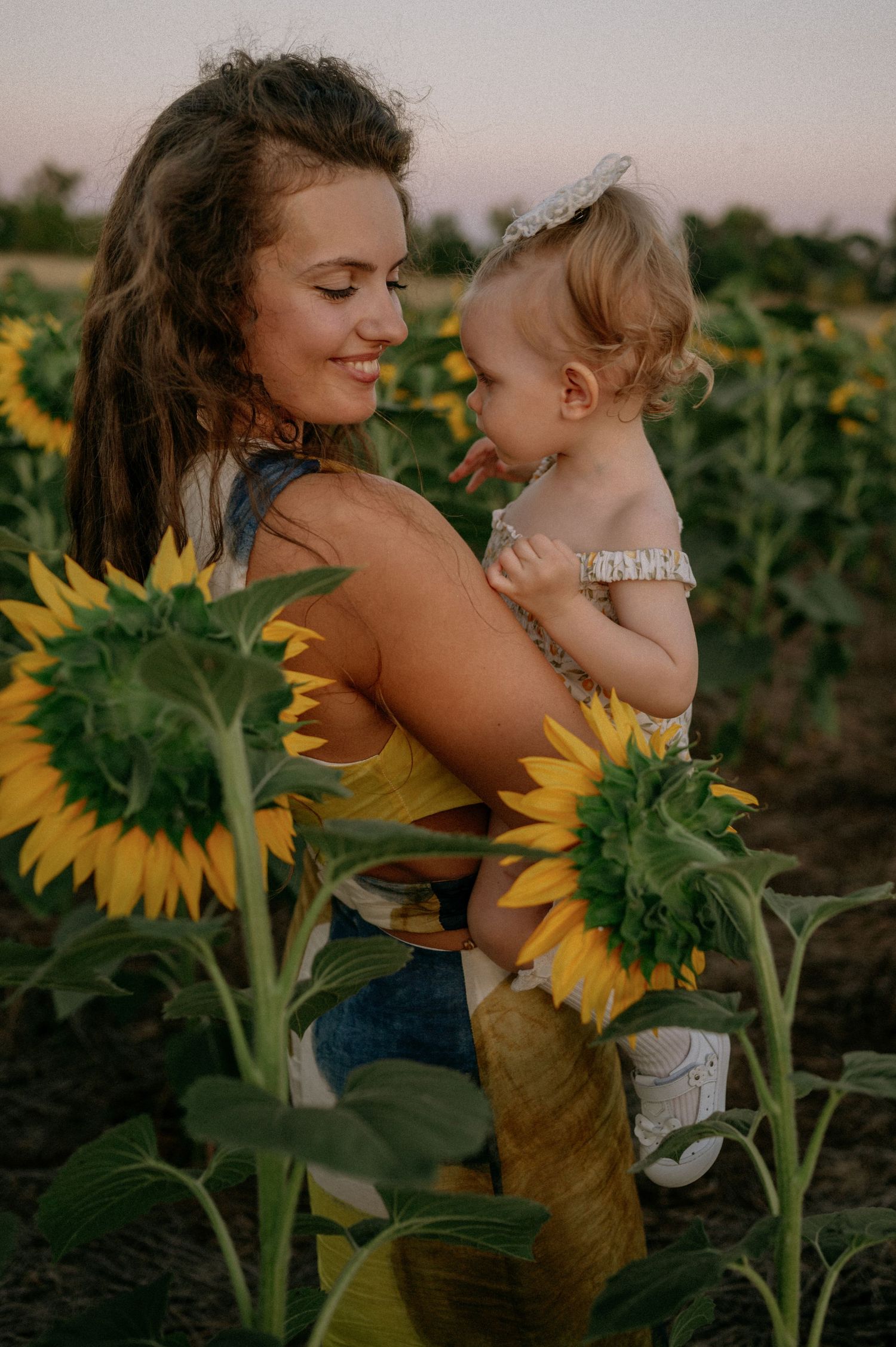 A smiling young mother holds her baby girl in a vibrant sunflower field at sunset.