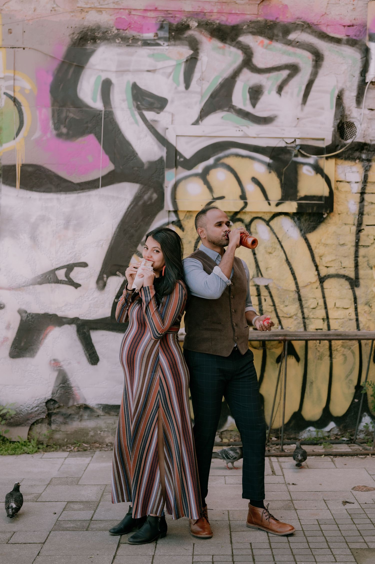 Couple posing back-to-back against a colorful graffiti wall, woman in striped dress, man in vest holding camera.