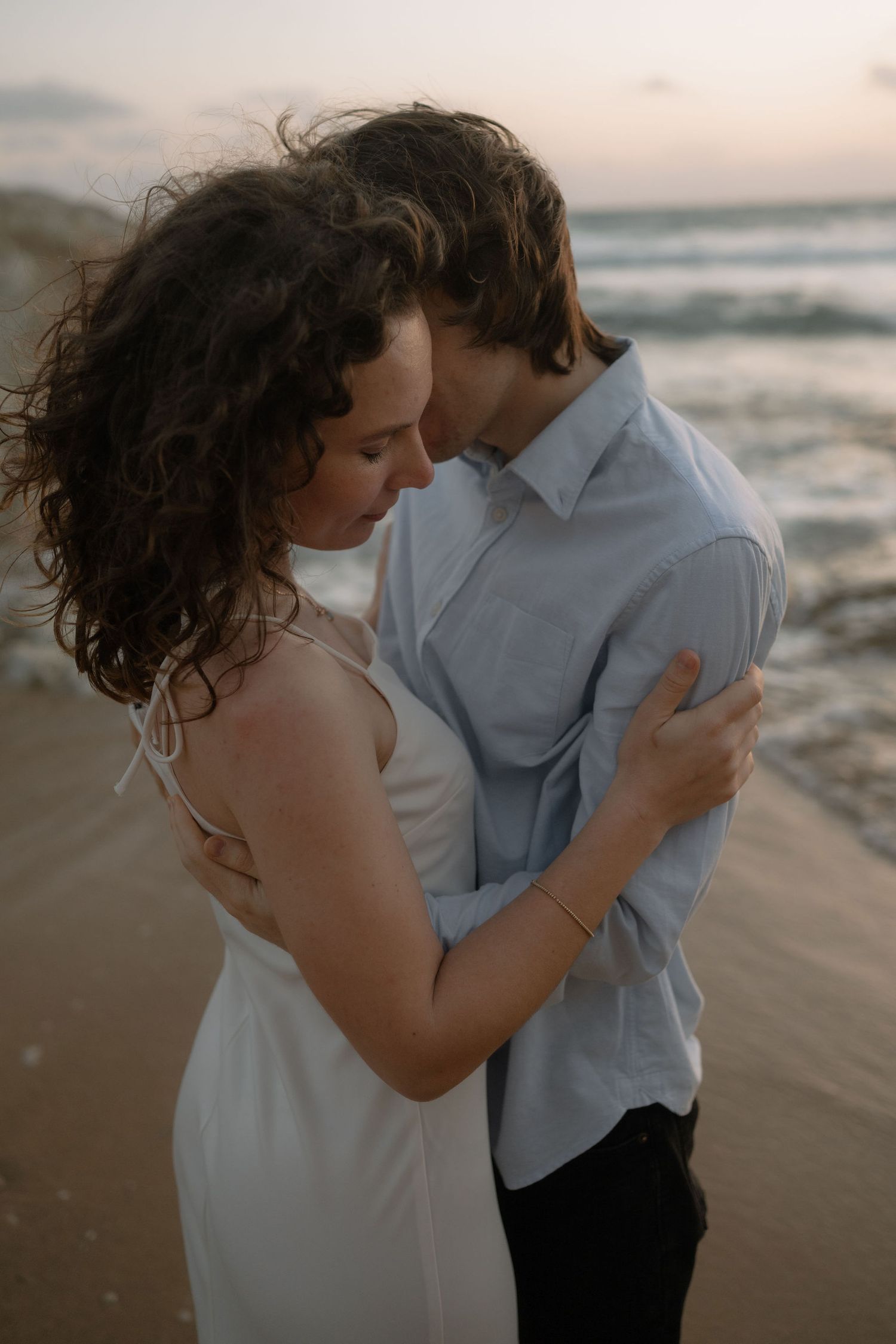 A couple embraces romantically on a beach at sunset, the woman in a white dress with curly hair blowing in the wind.