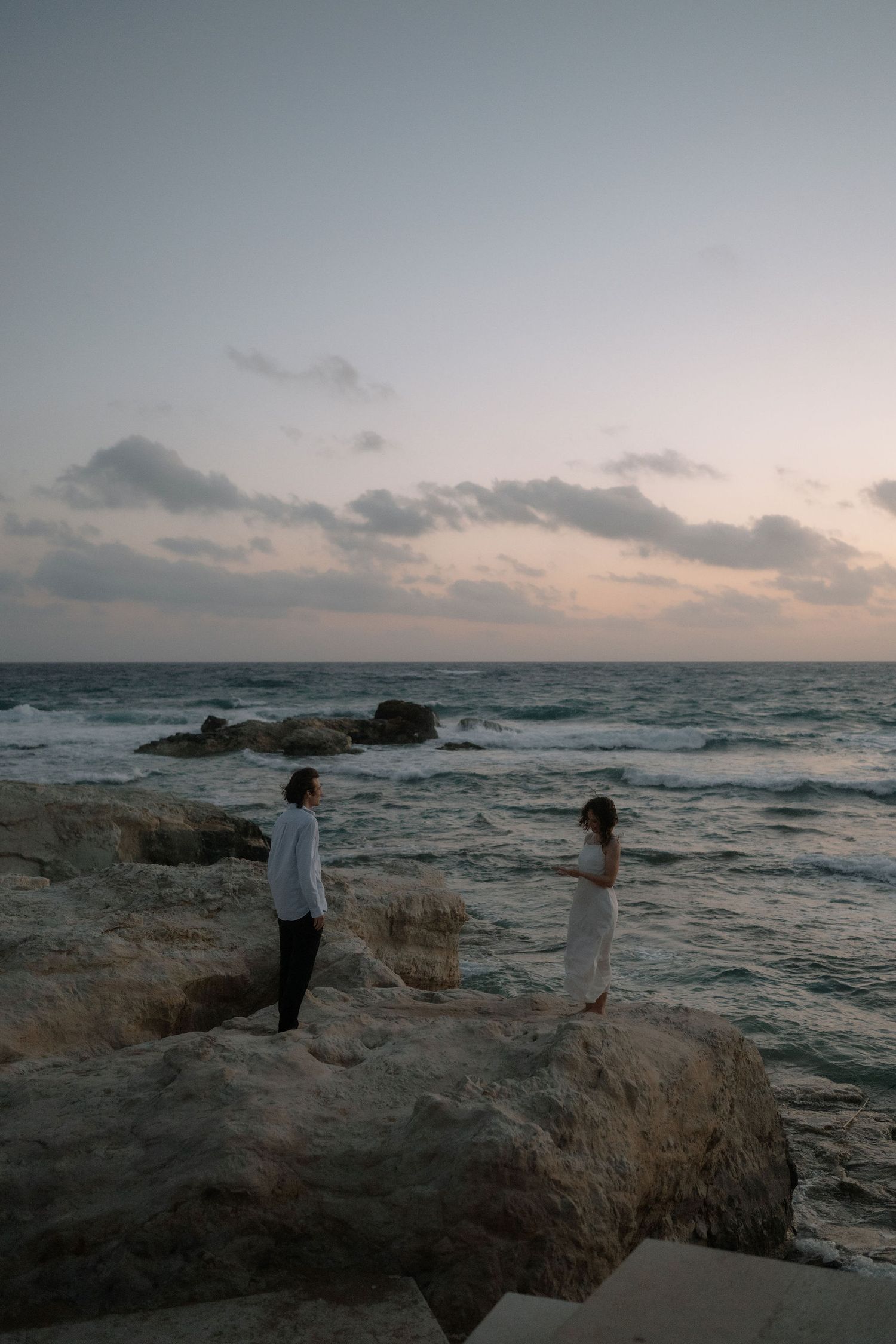 Two people stand on rocky shoreline at dusk, waves crashing behind them under a pastel cloudy sky.