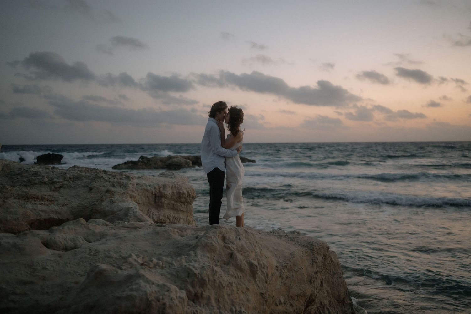 Couple embracing on rocky shoreline at sunset with ocean waves and dramatic sky in background.