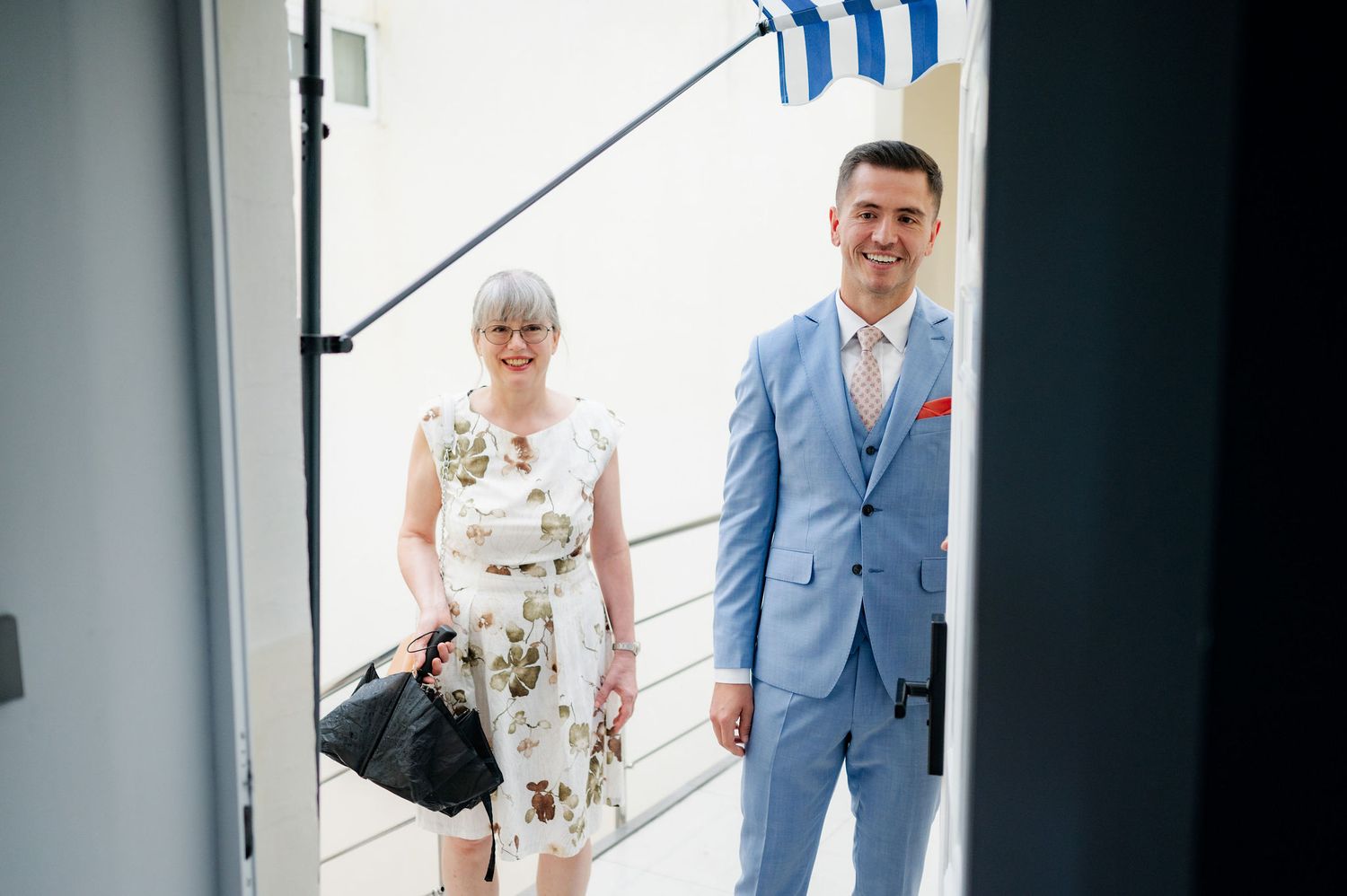 Two people stand in a bright doorway, with one wearing a floral dress and another in a light blue suit.