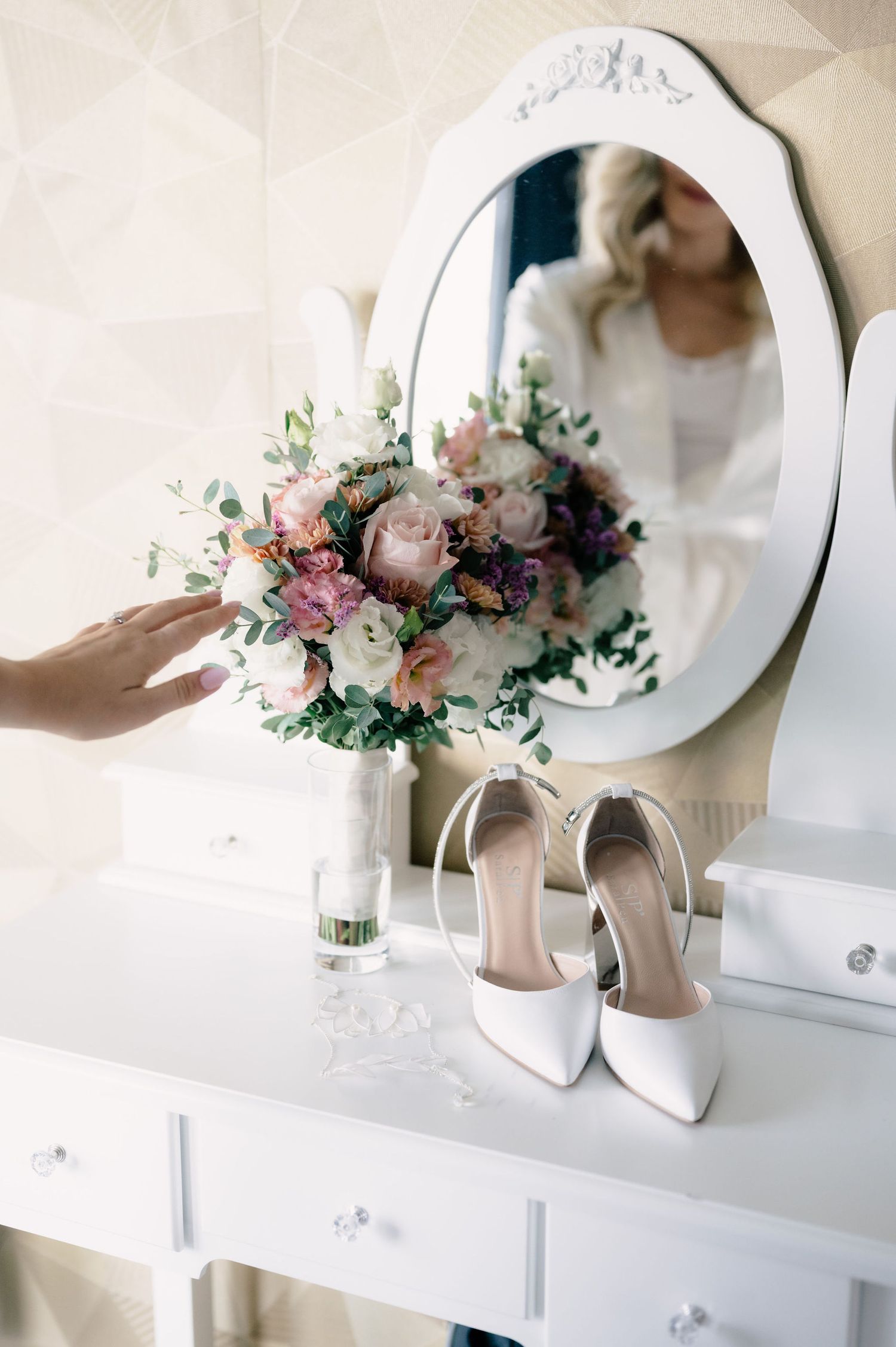 Pink and purple floral arrangement with roses sits elegantly next to beige high heel wedding shoes on white shelf.