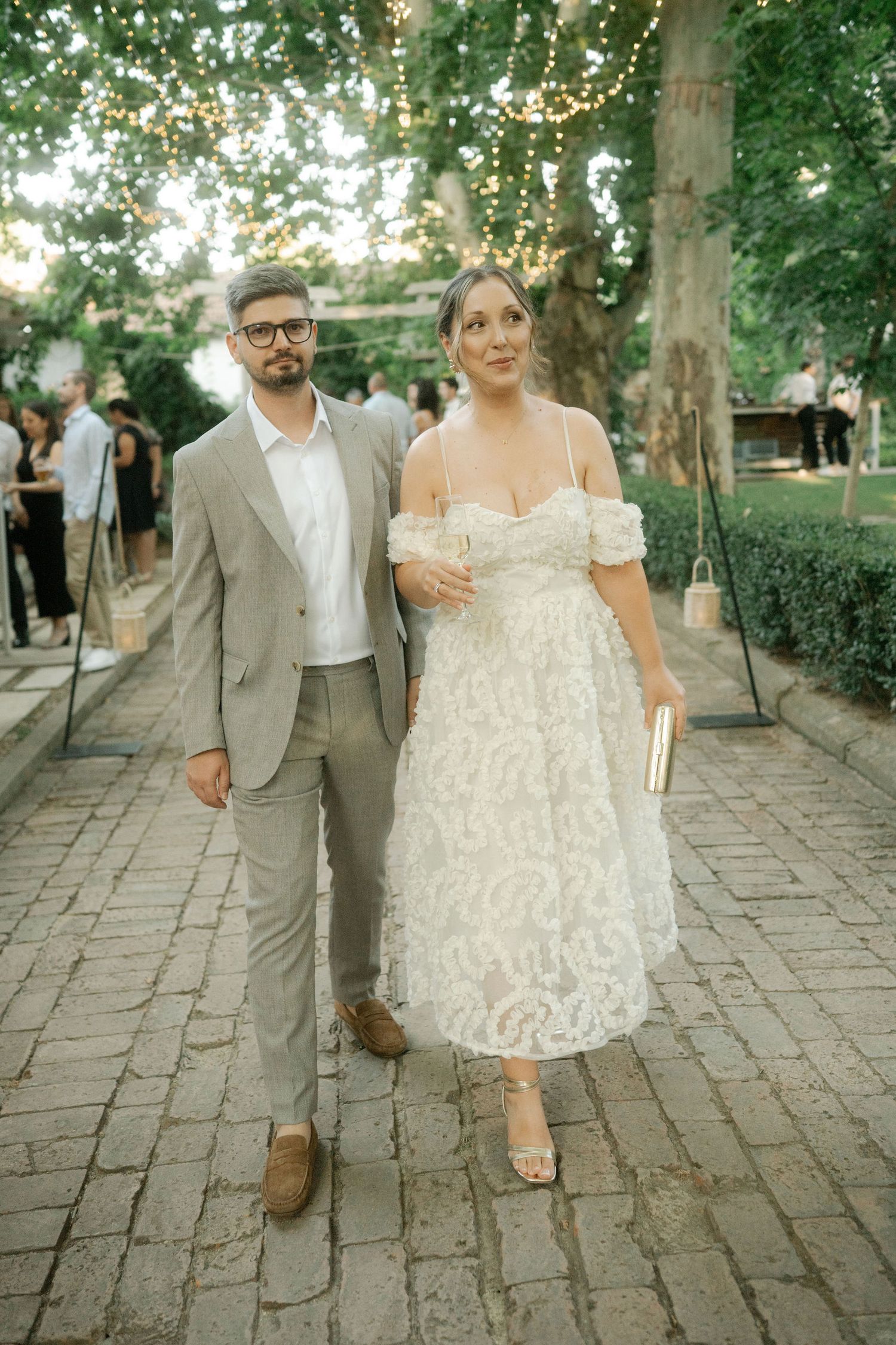 A couple walks hand in hand on a stone pathway during their outdoor wedding celebration at sunset.
