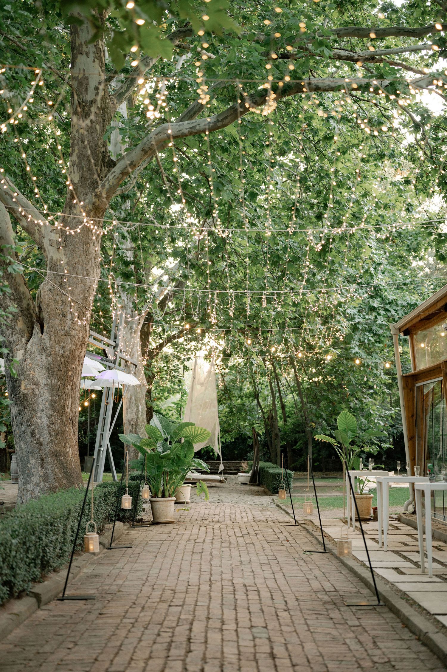 A serene brick-paved garden path winds through lush trees and greenery with wooden structures along the sides.