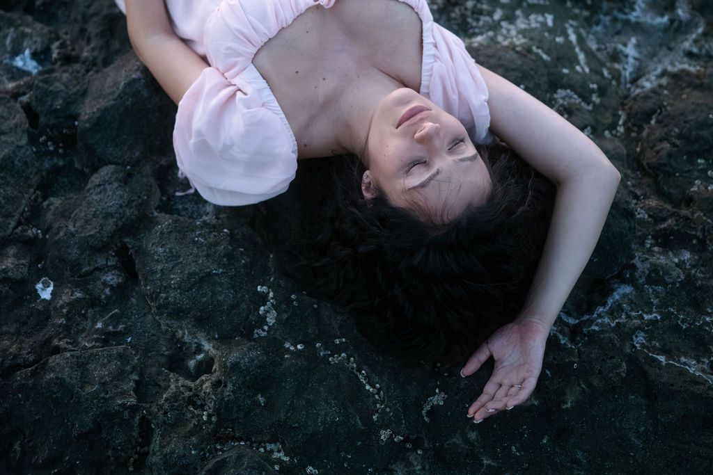 A person in white lies on dark rocks while posing for artistic outdoor portraits.