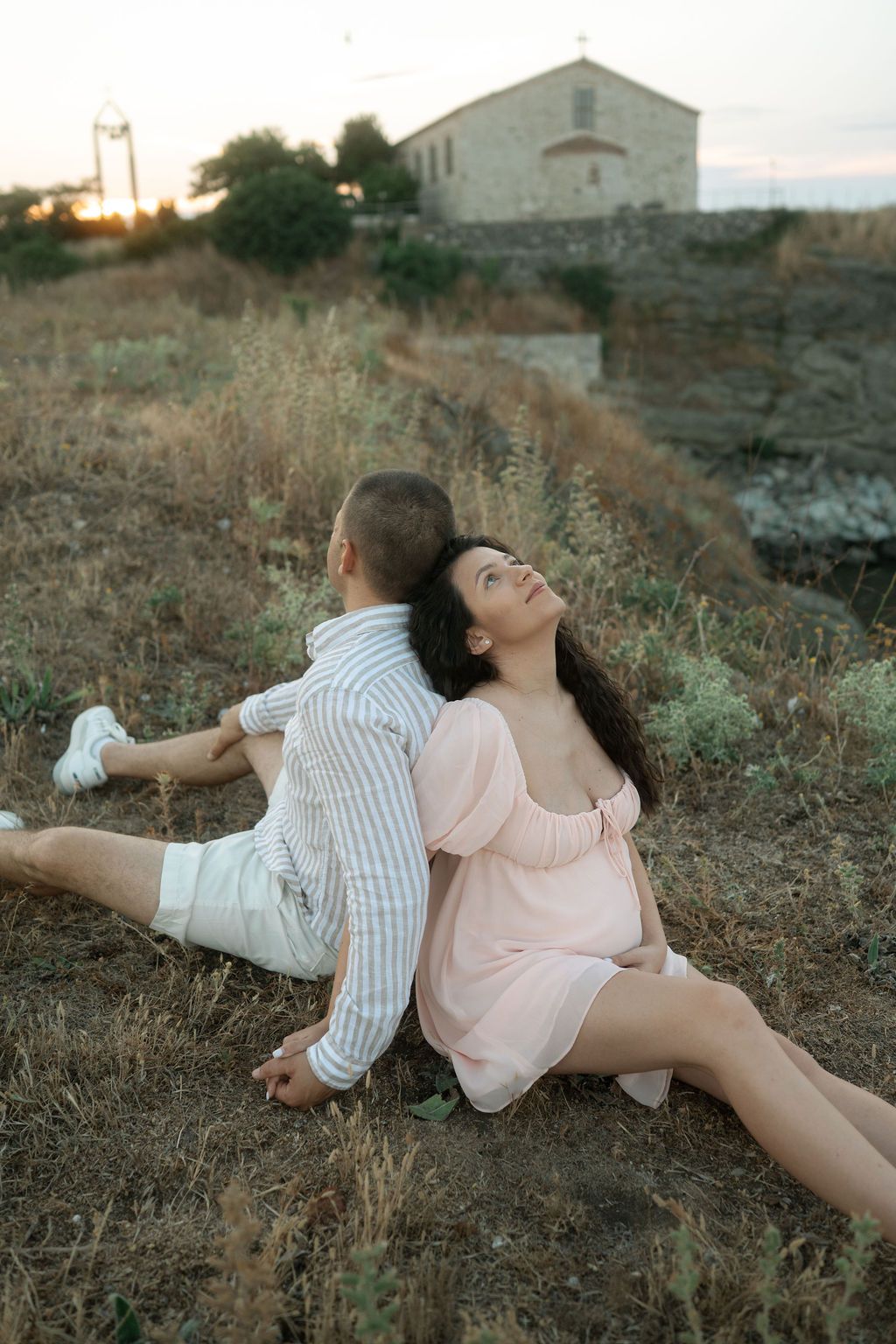 Two people in casual attire lying on grassy terrain near coastal buildings at dusk.