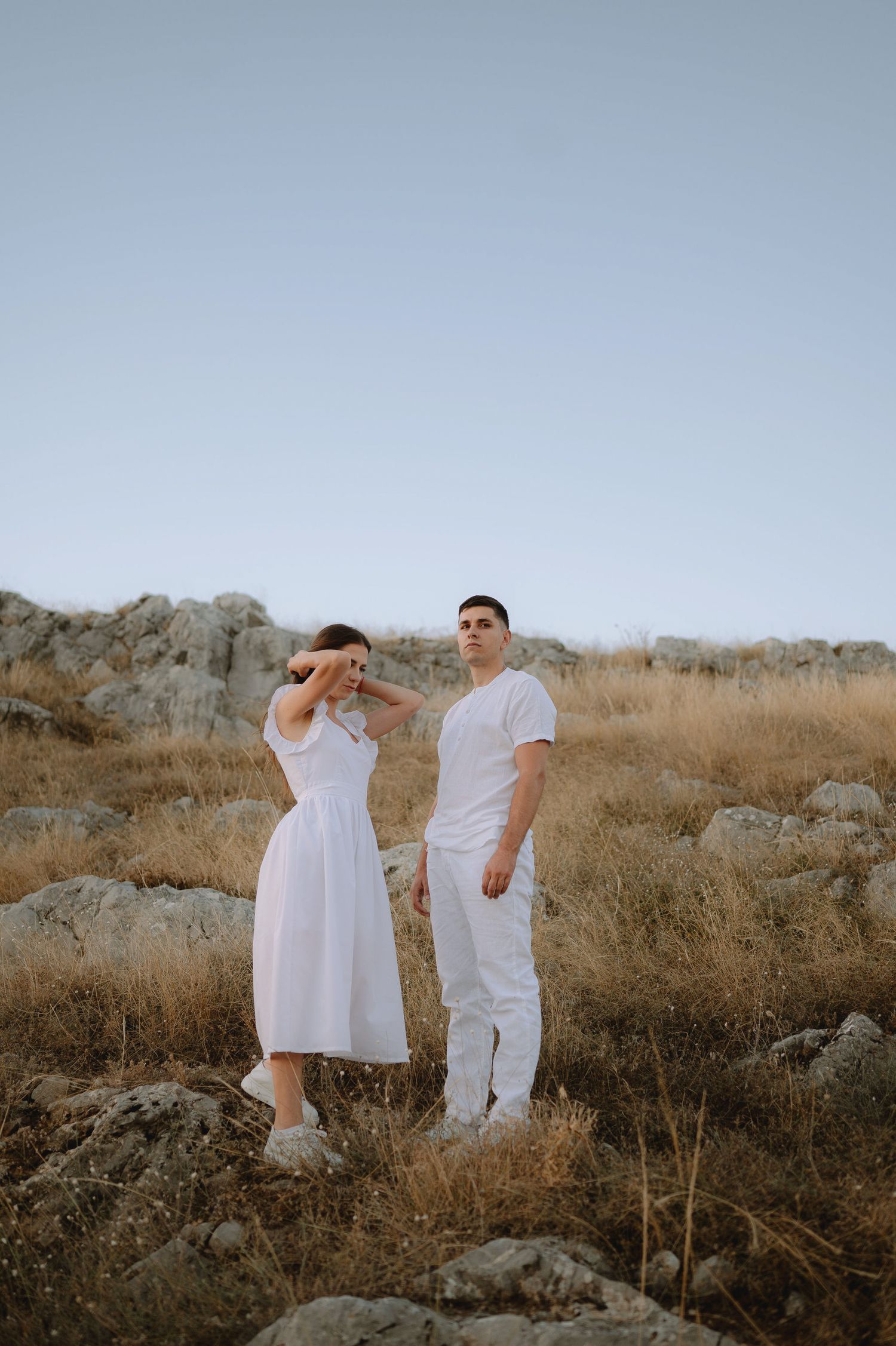 A couple in white outfits poses together in a desert landscape with dried brush and rocks at sunset.