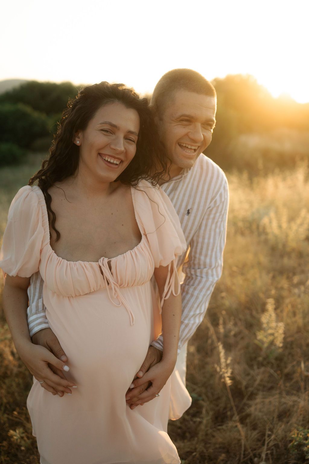 A couple embraces in a sunlit field during golden hour, wearing light colored clothing and sharing intimate moments.