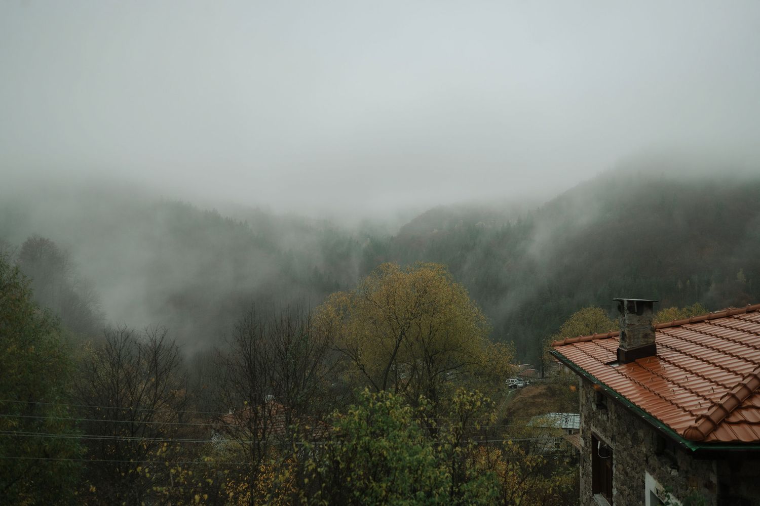 Misty mountain landscape with fog rolling over trees and a red-tiled roof in the foreground during autumn.