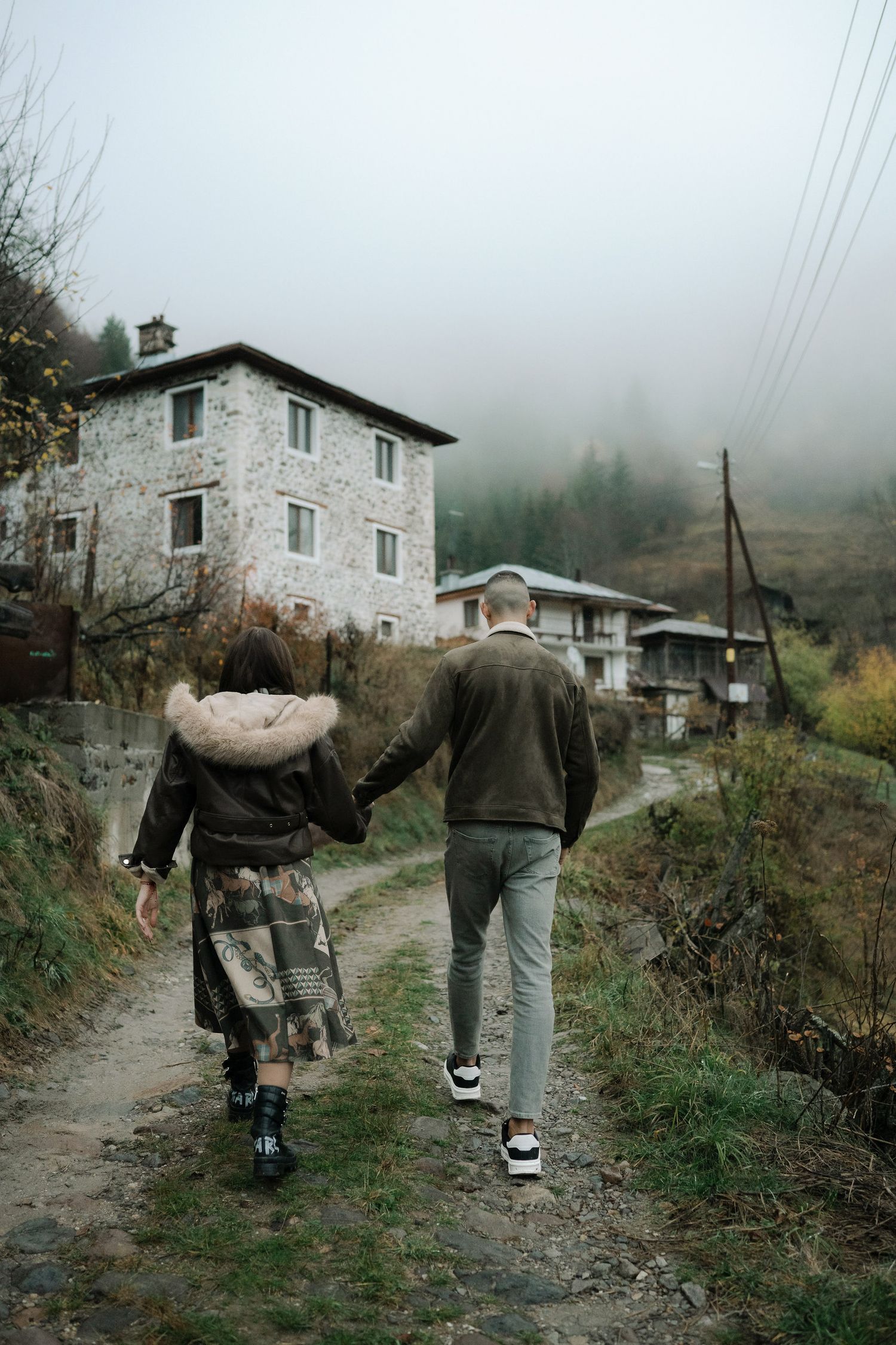 People walking along a rural mountain path carrying large sacks past stone buildings in misty weather.
