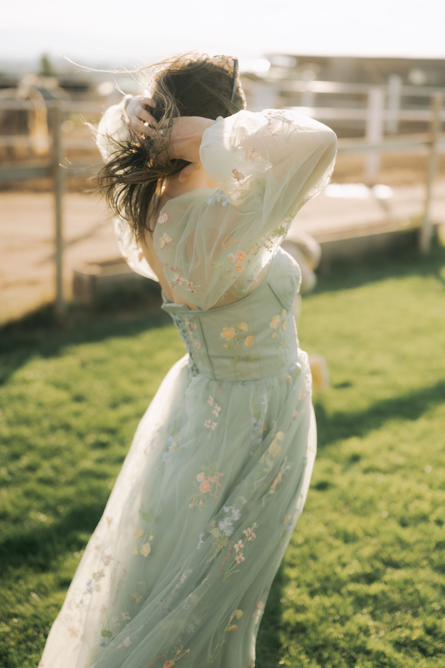 A person in a flowing mint green dress twirls gracefully in a sunlit field with wooden fencing in the background.