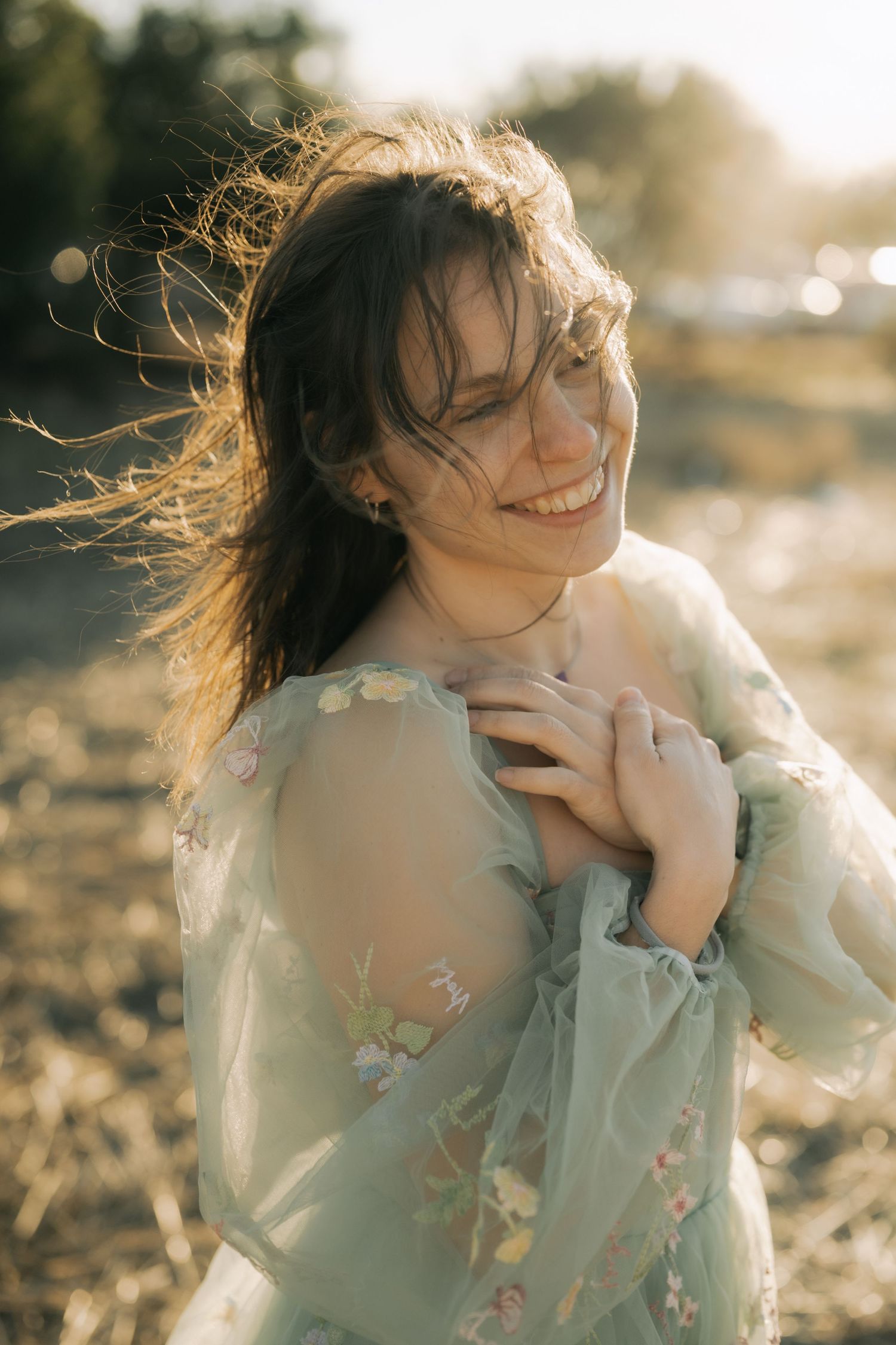 A person in a mint green floral dress laughs joyfully while their hair blows in the sunlit breeze.