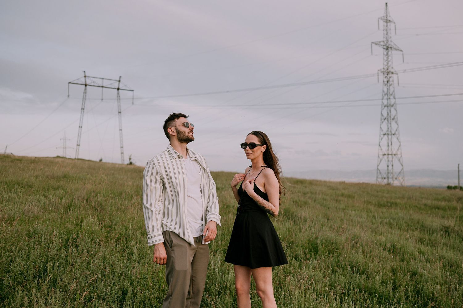Two people walk together through a green field with electrical transmission towers in the background on an overcast day.