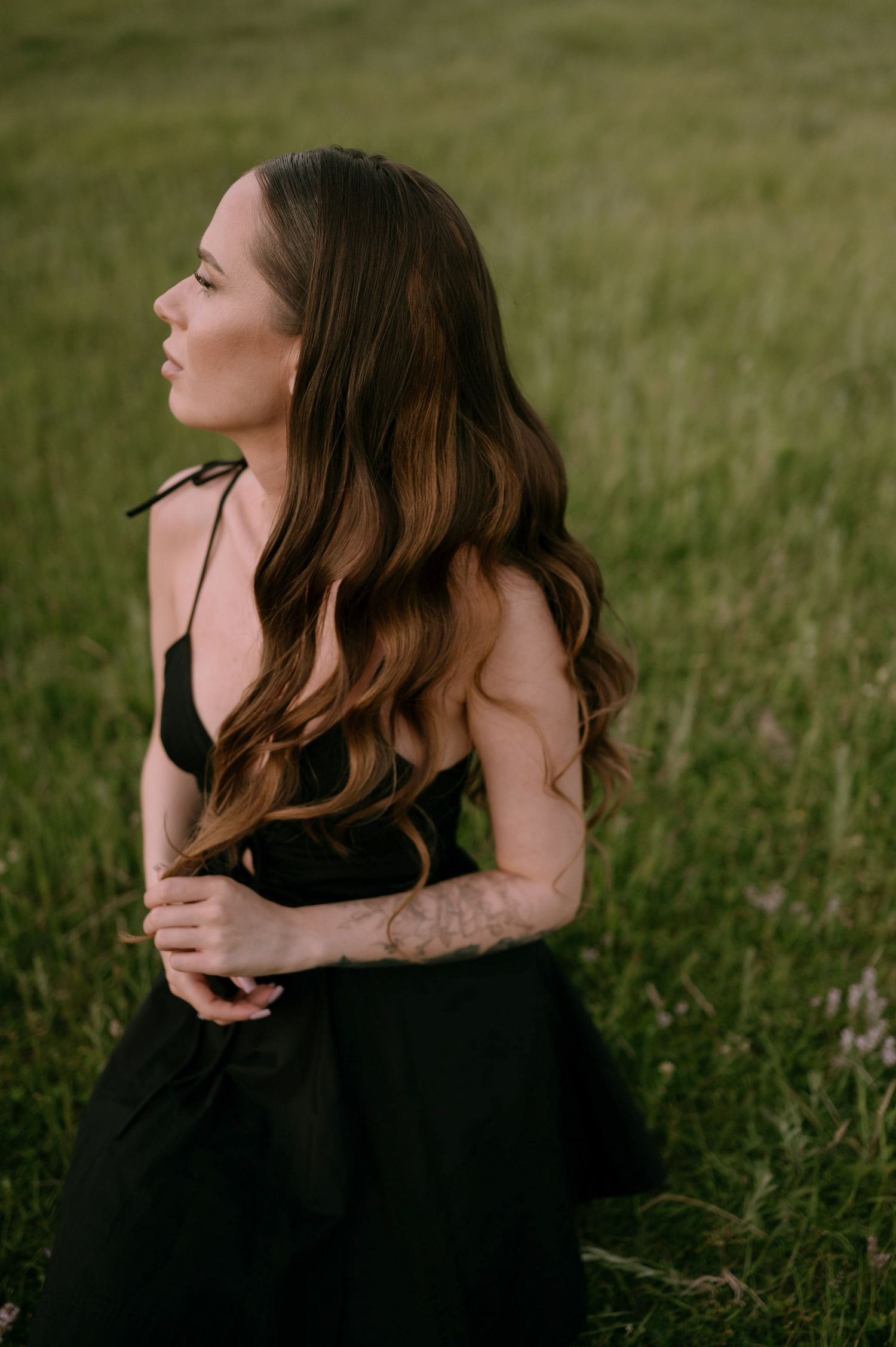 A woman in a black dress poses gracefully in a grassy field during a moody outdoor photoshoot.
