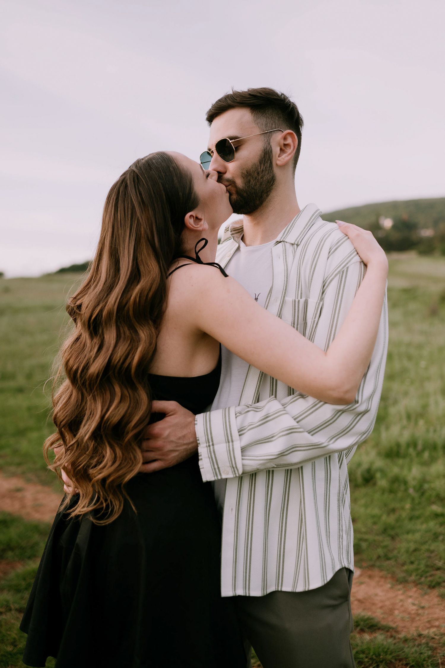 A romantic couple embraces on a grassy hillside at sunset, with her in a black dress and him in a striped shirt.