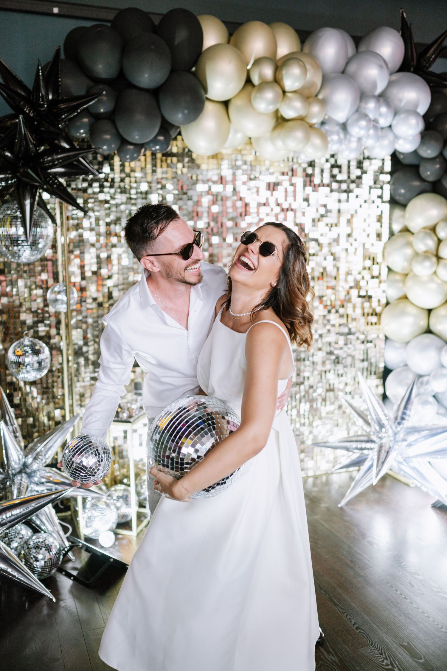 A couple in white outfits dances and laughs together under a ceiling decorated with silver disco balls and balloons.