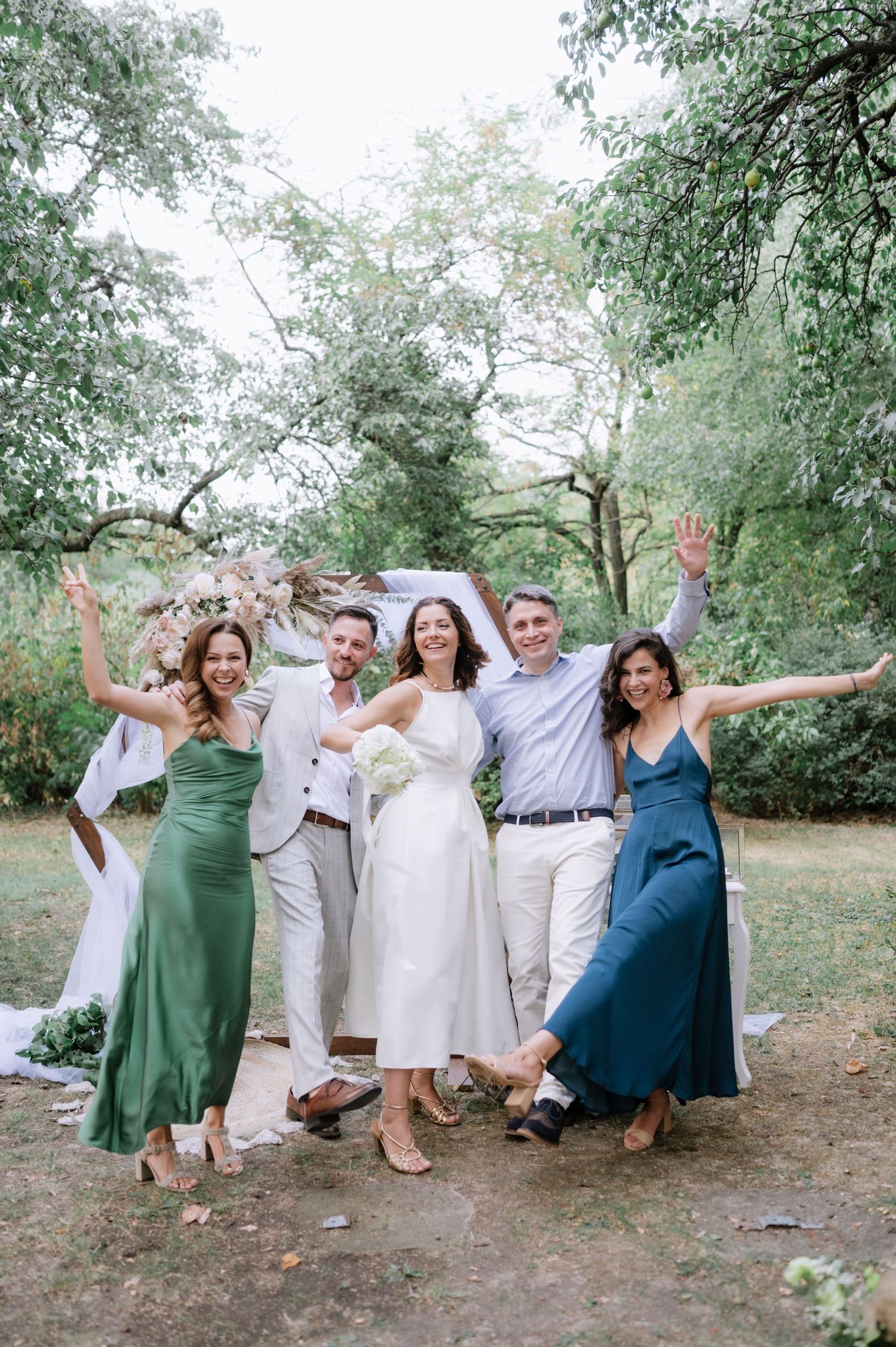 A group of wedding guests strike playful poses while sitting on a fallen tree trunk in a natural outdoor setting.