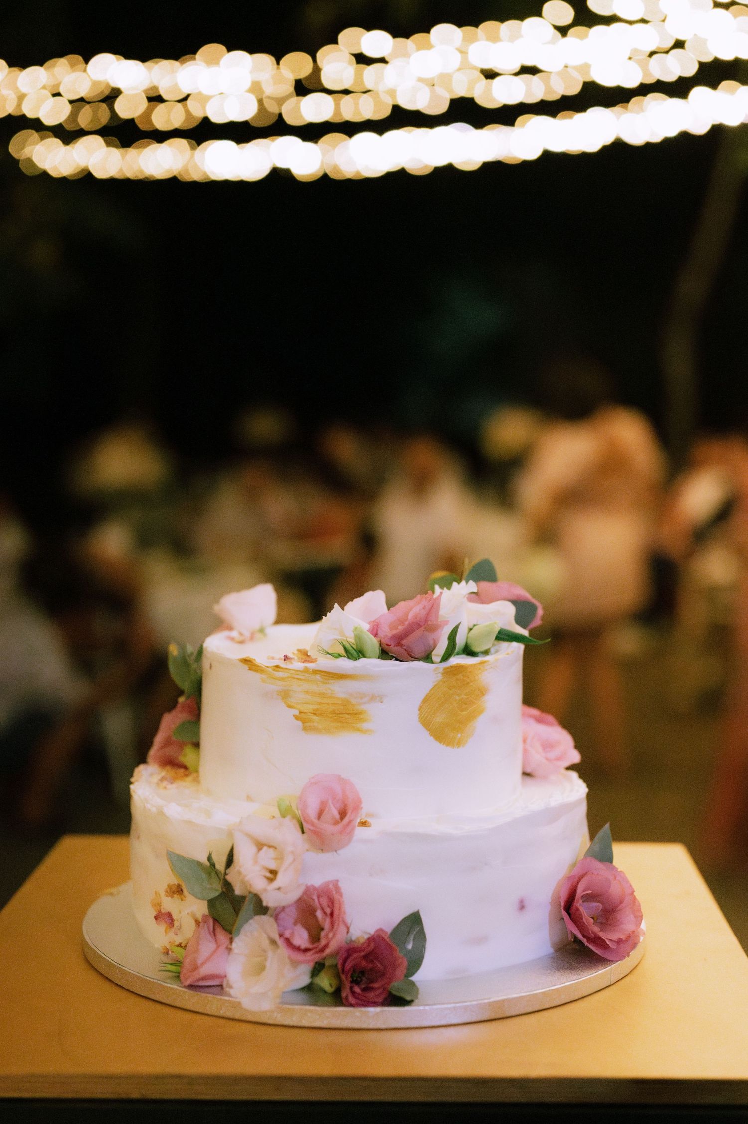 Two-tiered white wedding cake decorated with pink roses and gold accents under string lights at evening reception.