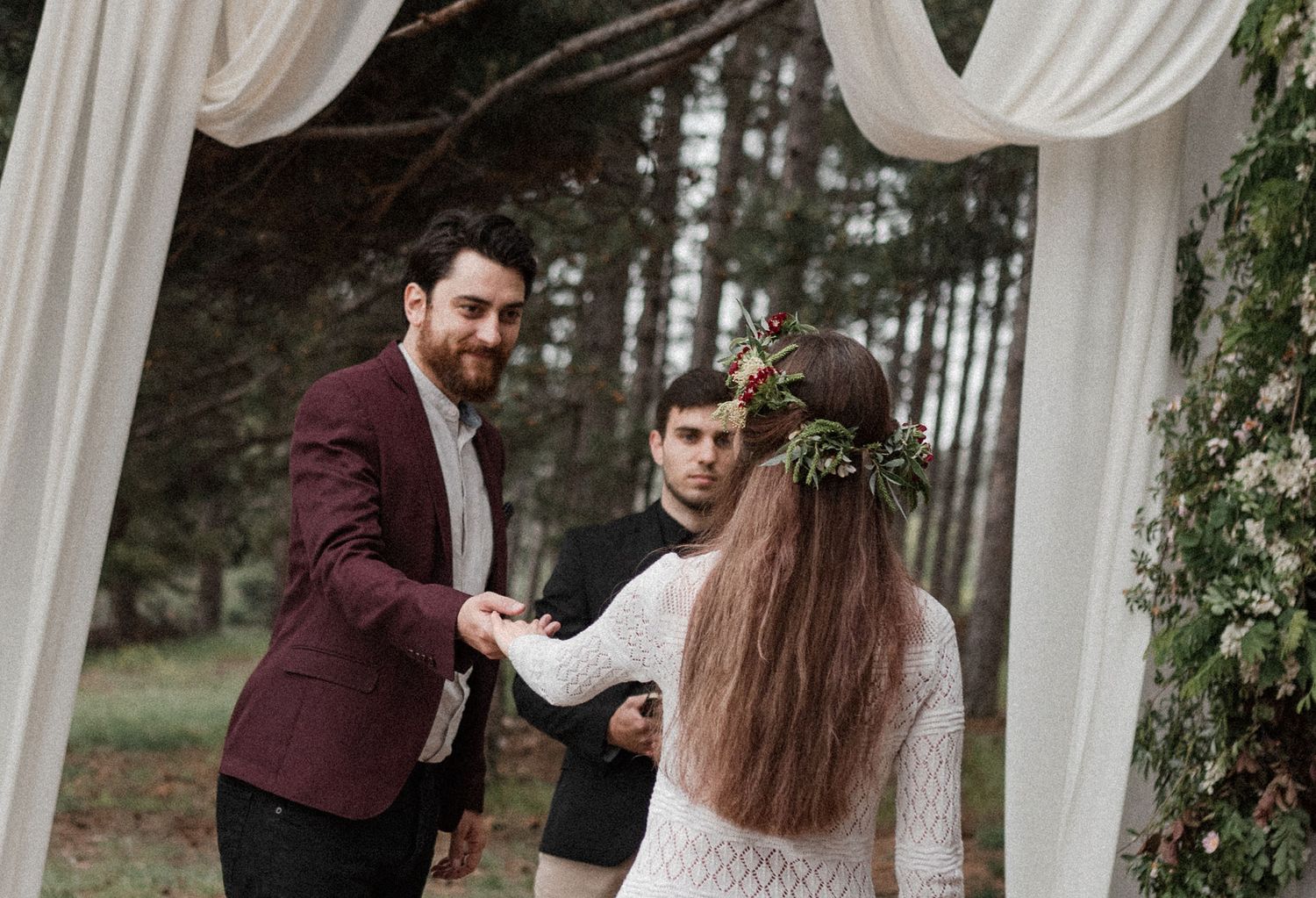 A couple exchanges vows during an outdoor wedding ceremony under a white draped gazebo surrounded by pine trees.