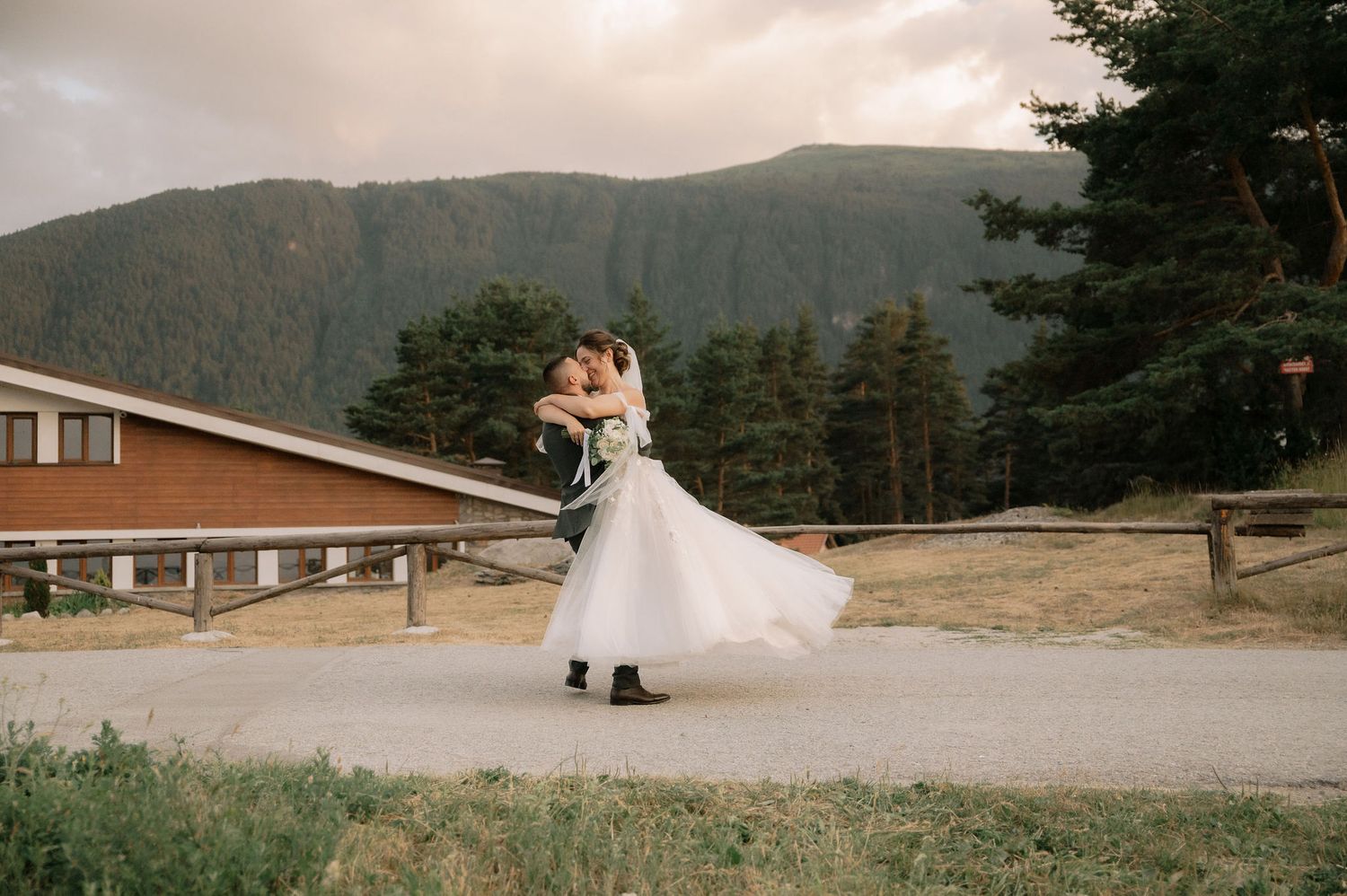 A couple in wedding attire stands on a scenic mountain overlook with evergreen trees and mountains in the background.
