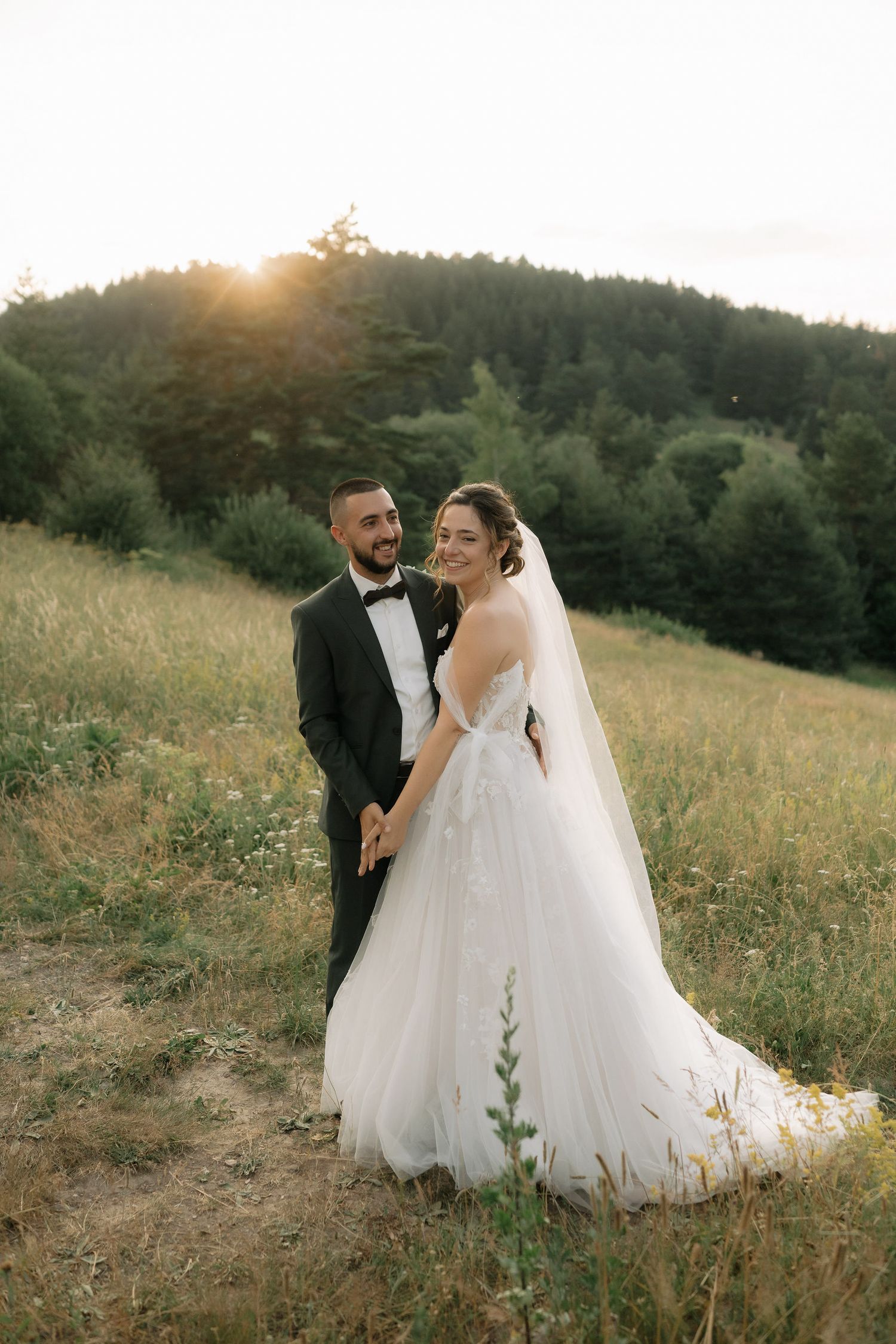 A couple shares a romantic moment in their wedding attire on a sunlit hillside with wildflowers at golden hour.