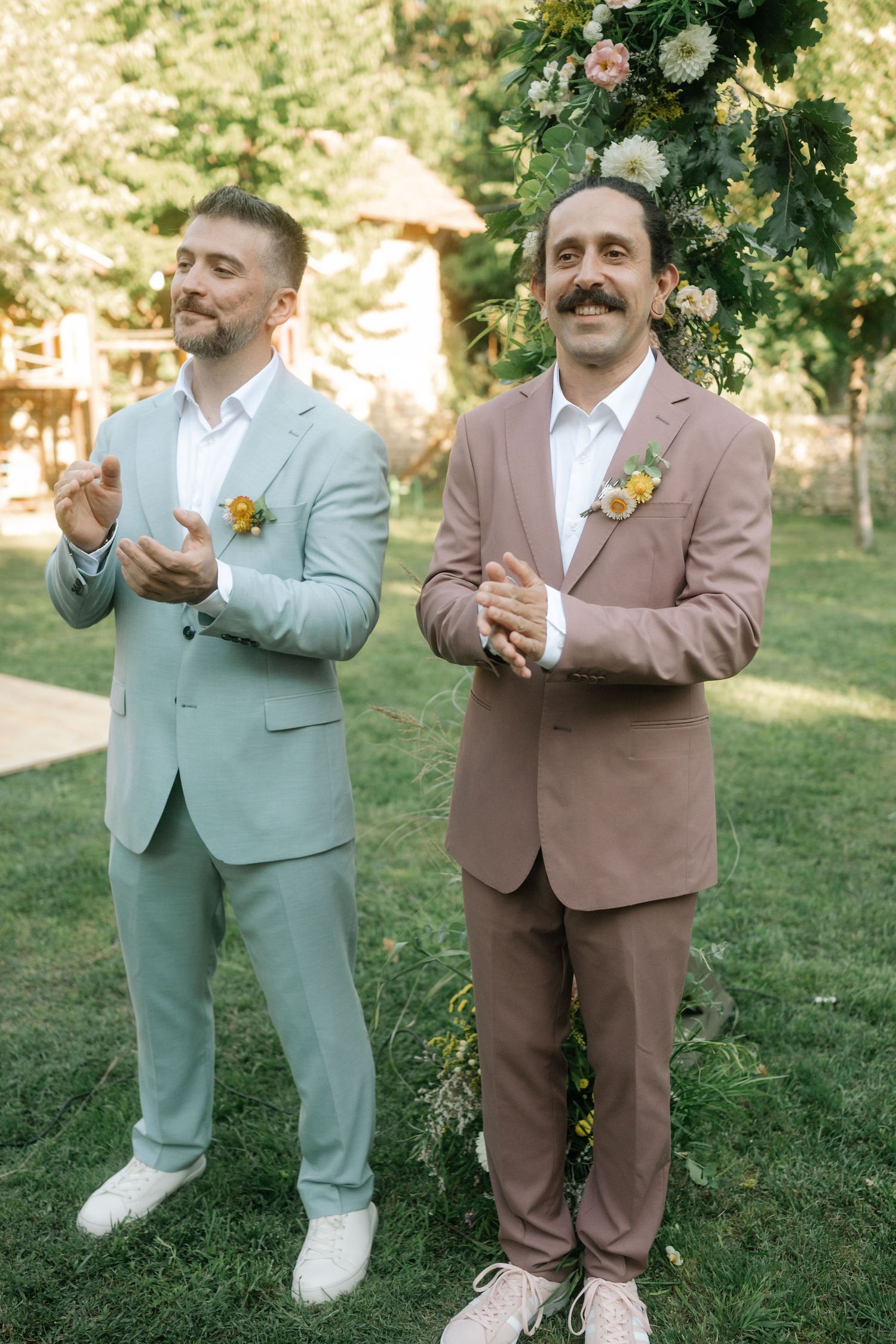 Two grooms in pastel suits stand together on a lawn during an outdoor wedding celebration.