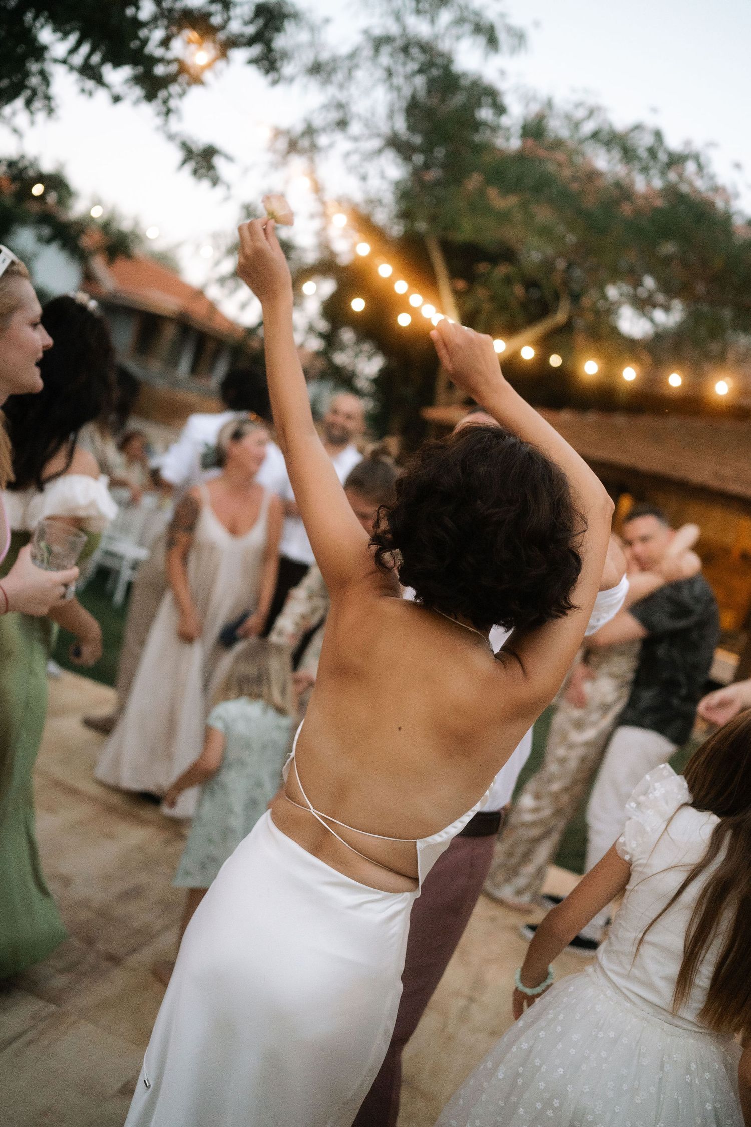 People dancing and celebrating at an outdoor wedding reception under string lights at dusk.